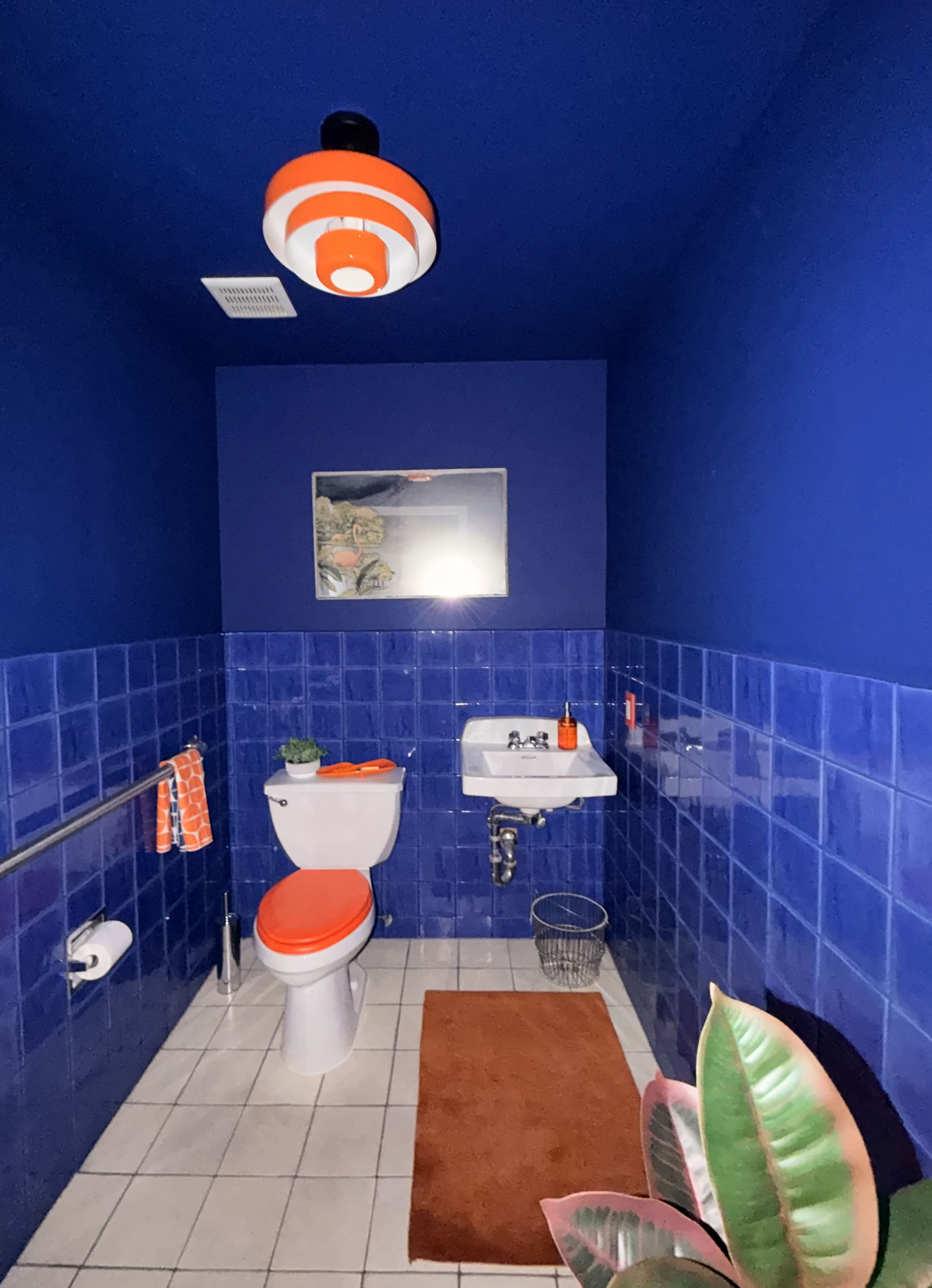 A small bathroom features blue walls, blue tiled accents, a white toilet, a wall-mounted sink, an orange striped light fixture, and decorative plants.