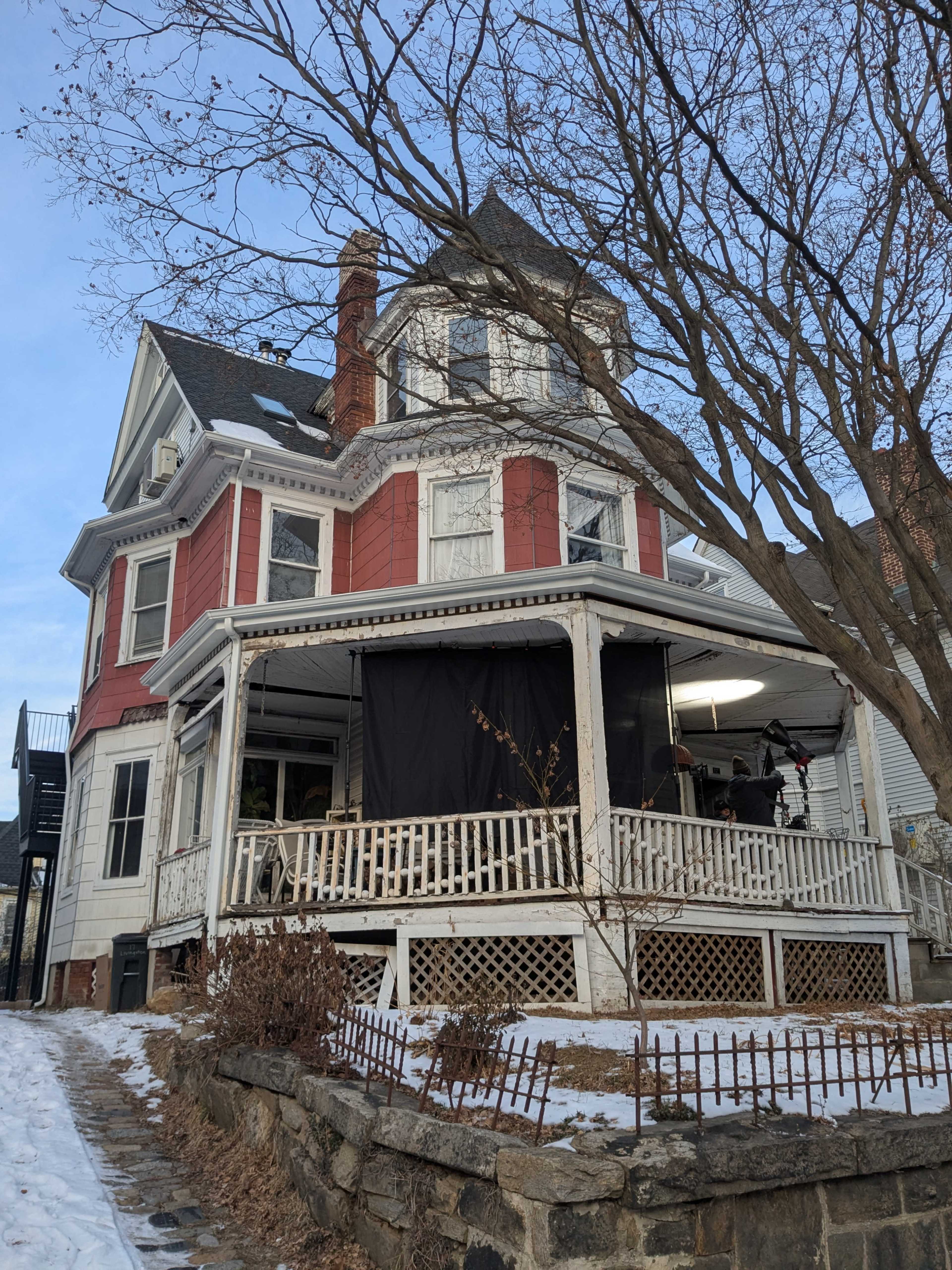 A Victorian-style house with a distinctive turret features a wraparound porch and is partially covered by a black tarp, surrounded by a snowy landscape.