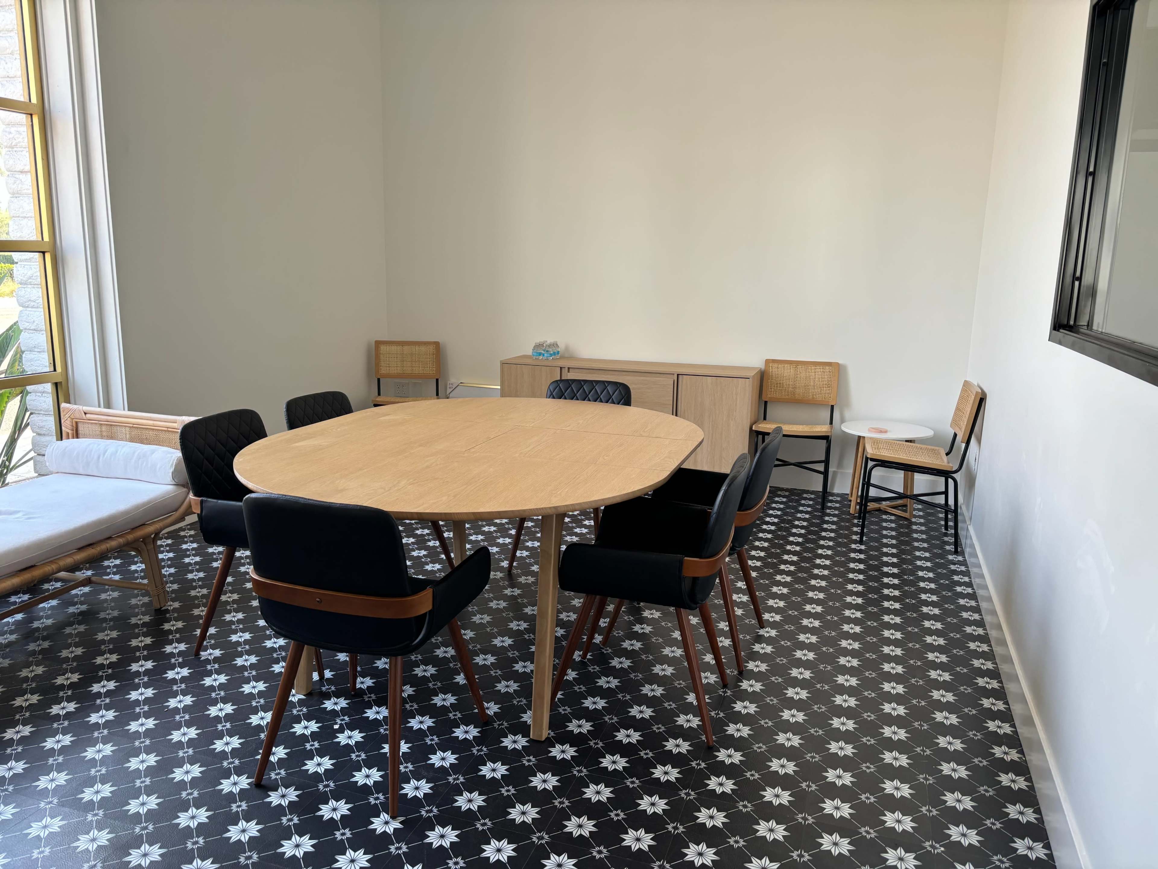 A round wooden table surrounded by black and wooden chairs is set in a minimally decorated room with patterned flooring.