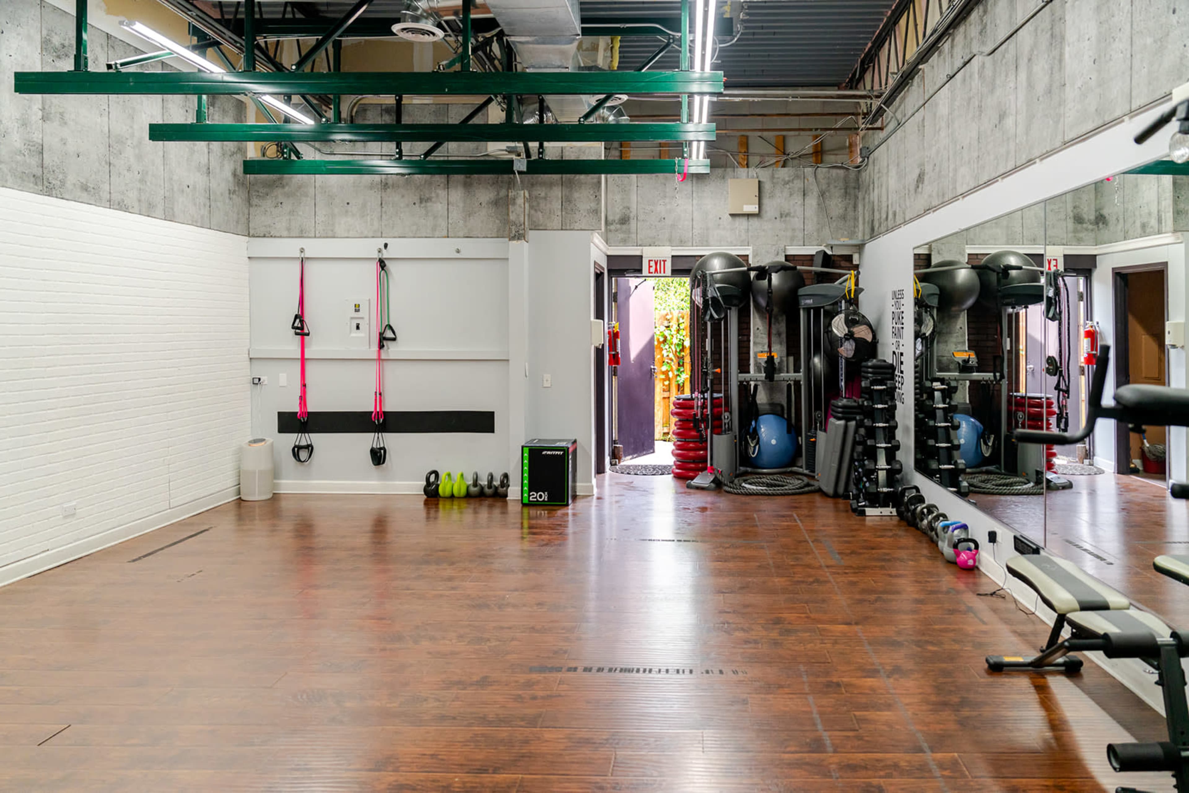 The image shows an empty gym space with fitness equipment neatly arranged against the walls and a doorway leading to an outdoor area.