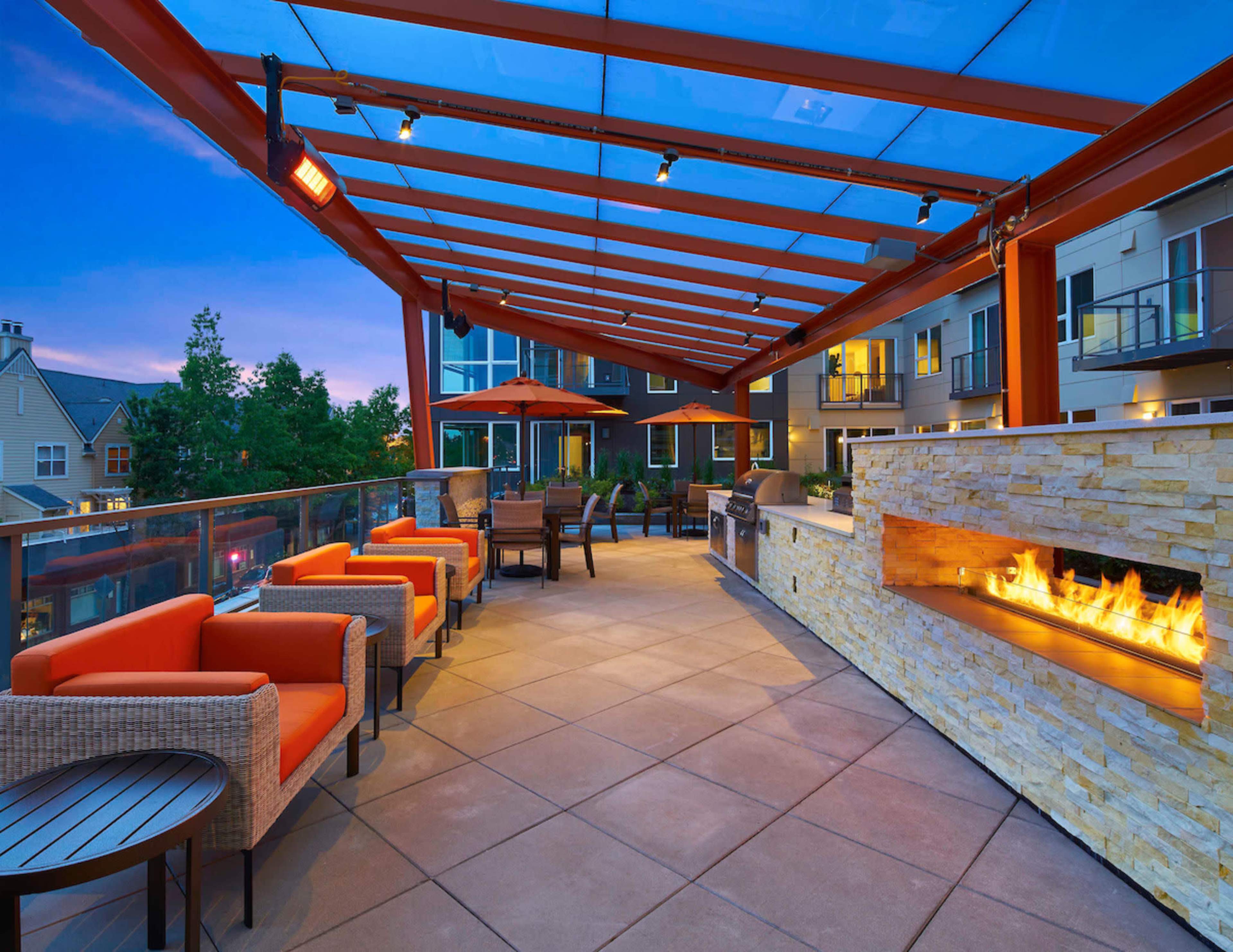 The image shows a modern outdoor seating area with orange chairs, a stone fireplace, and a grill under a red pergola.
