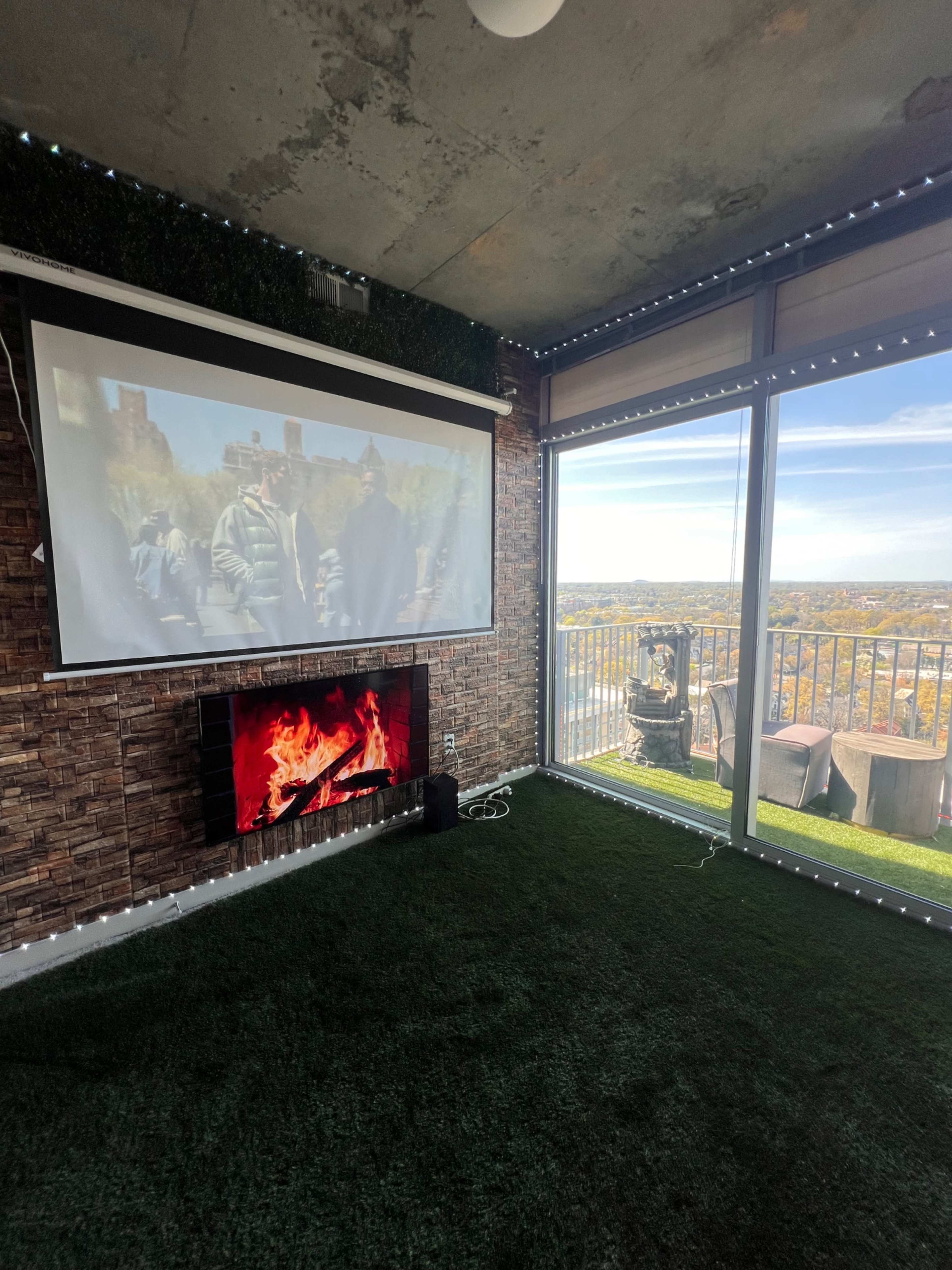 The room features a large projection screen displaying a scene from a film, with a modern faux fireplace beneath it and a view of the outdoors through a large glass window.