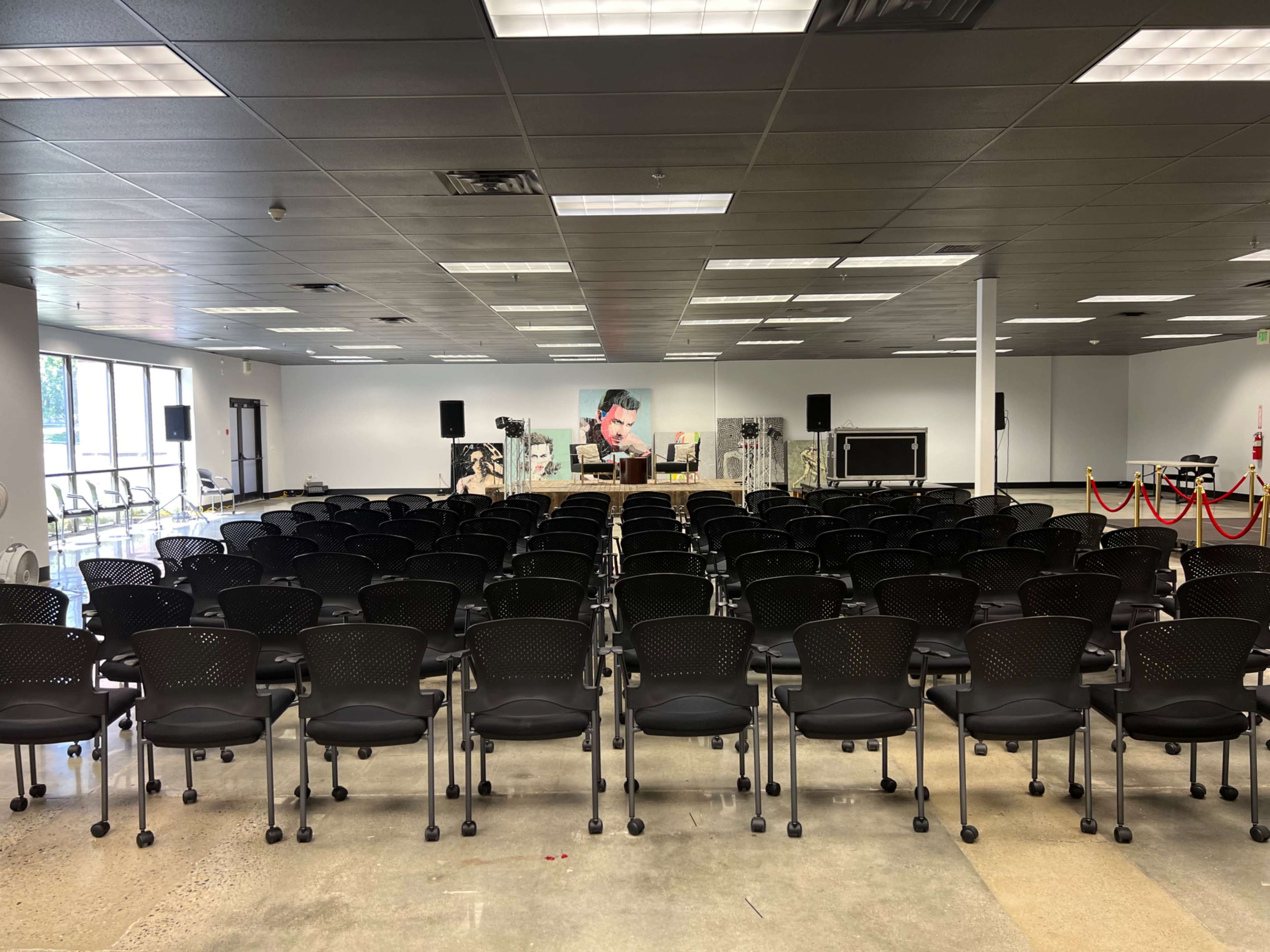 A spacious event room is set up with rows of black chairs facing a stage adorned with a backdrop and two armchairs.