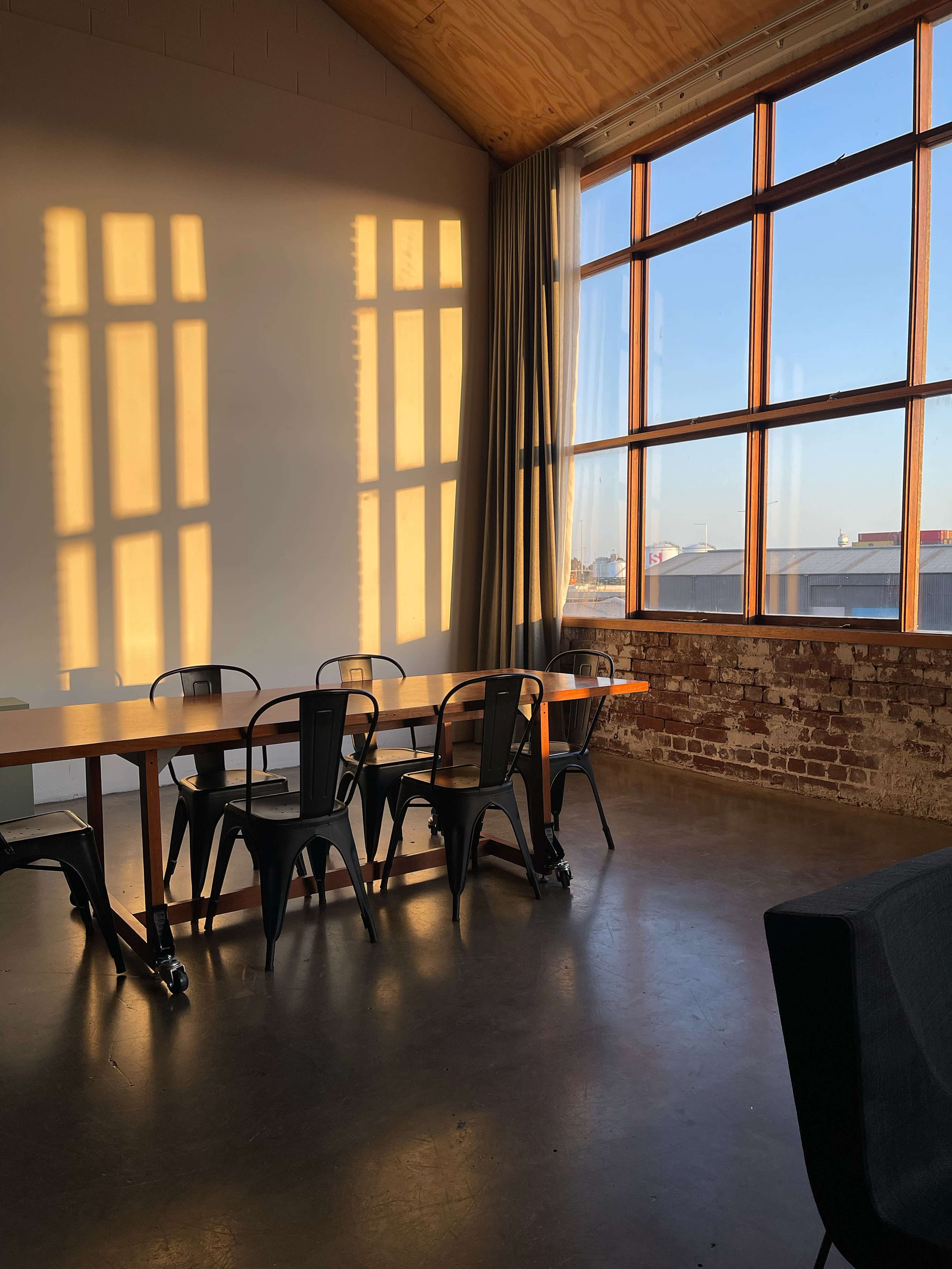A long wooden table with metal chairs is positioned near large windows, casting shadows on the wall as sunlight streams into the room.