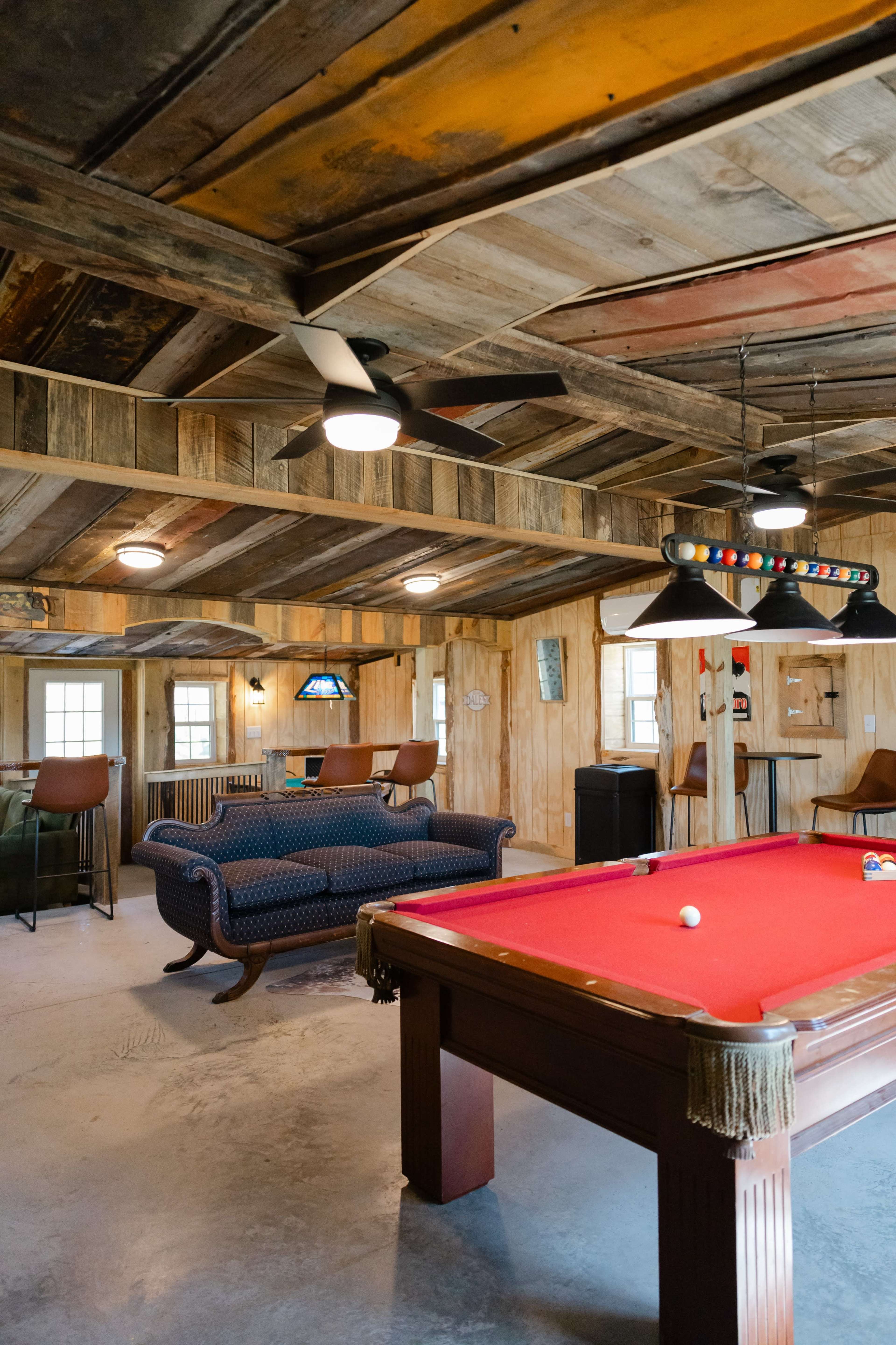 The image shows a rustic game room featuring a billiard table, a blue sofa, and wooden beams on the ceiling.