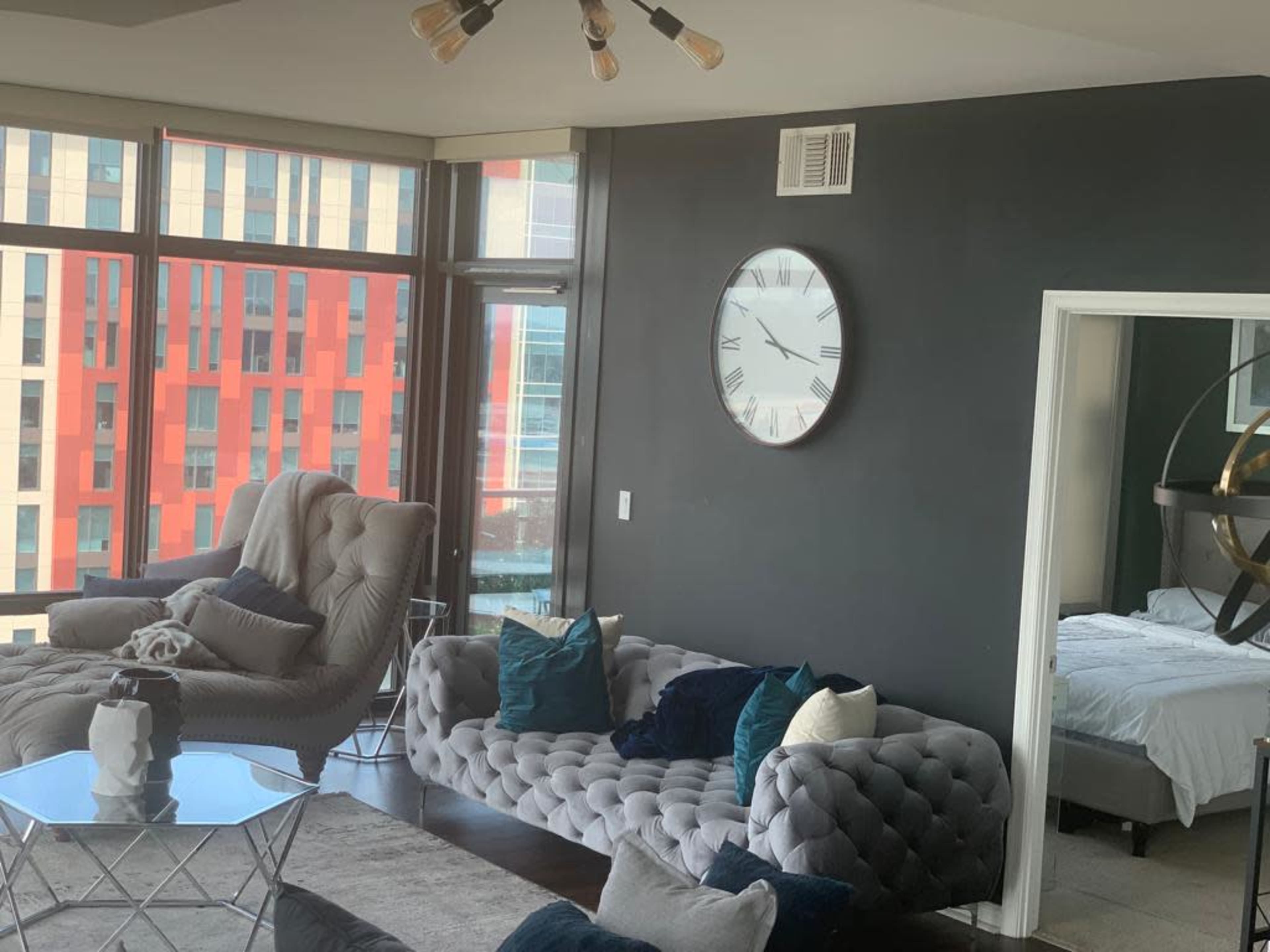 A modern living room with large windows, a circular clock on the wall, and a cozy seating area featuring a gray sofa and an armchair.