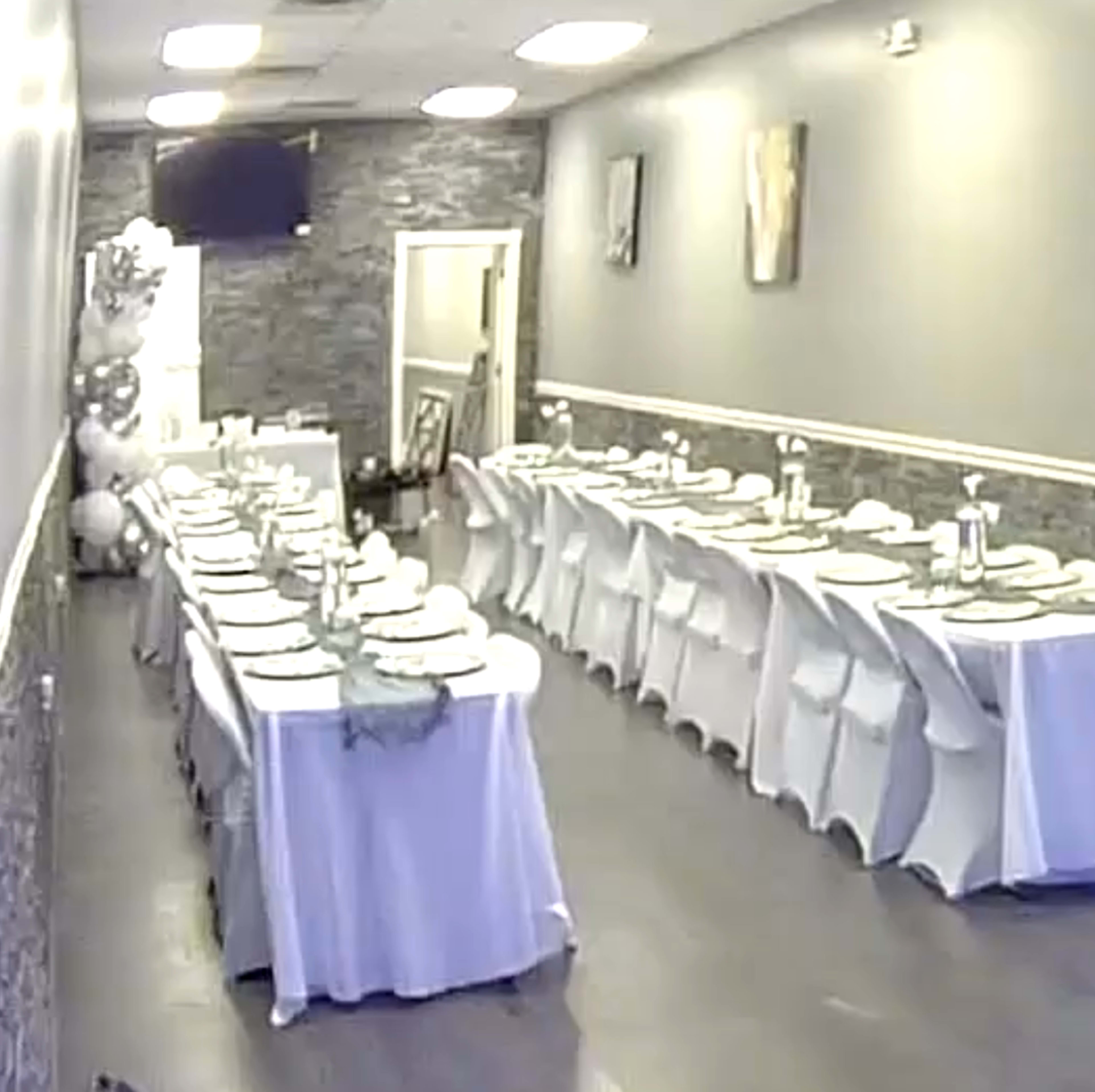 The image shows a banquet hall setup with multiple tables covered in white tablecloths arranged in a long line, prepared with dishware for a gathering.