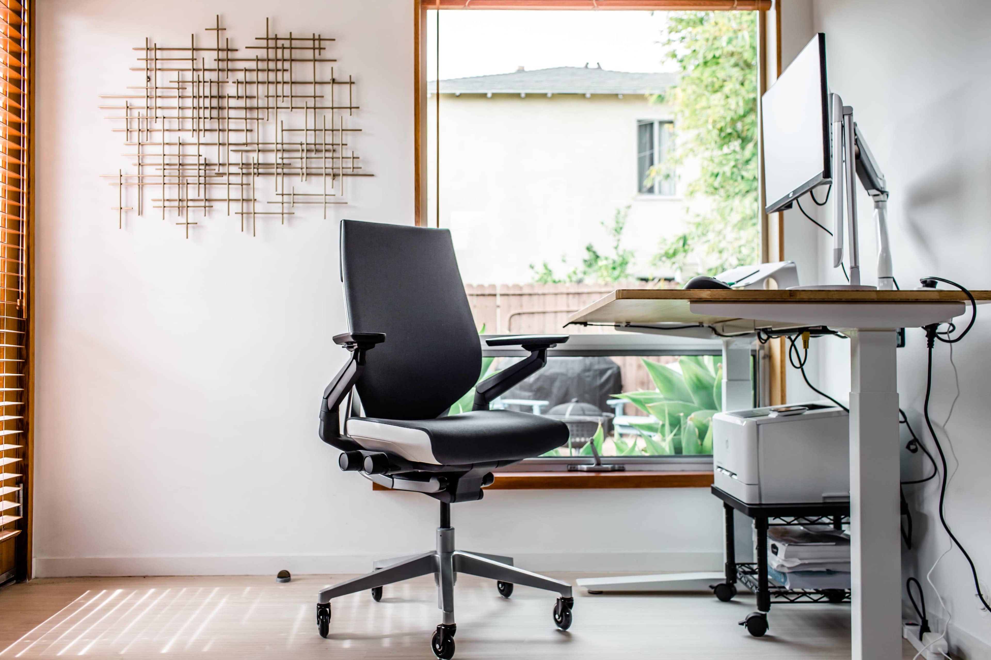 A black ergonomic chair is positioned in front of a desk with a computer monitor, beside a large window with wooden blinds.
