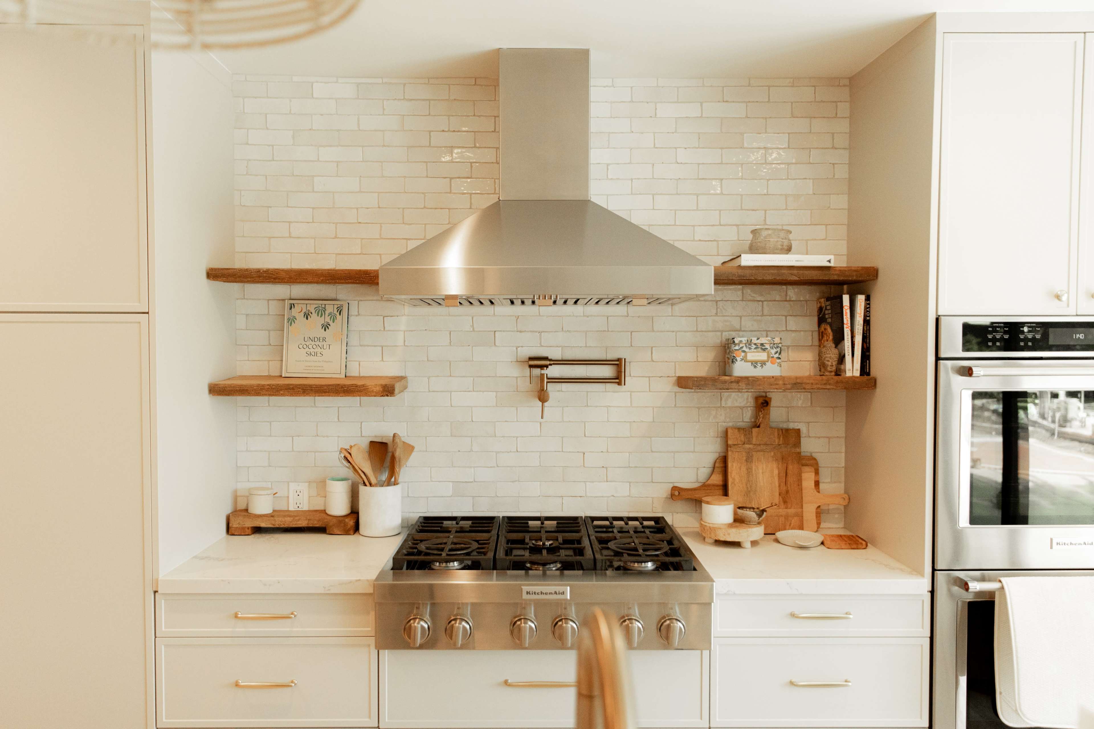 The image shows a modern kitchen featuring a stainless steel range hood above a six-burner gas stove, flanked by wooden shelves with decorative items against a tiled backsplash.