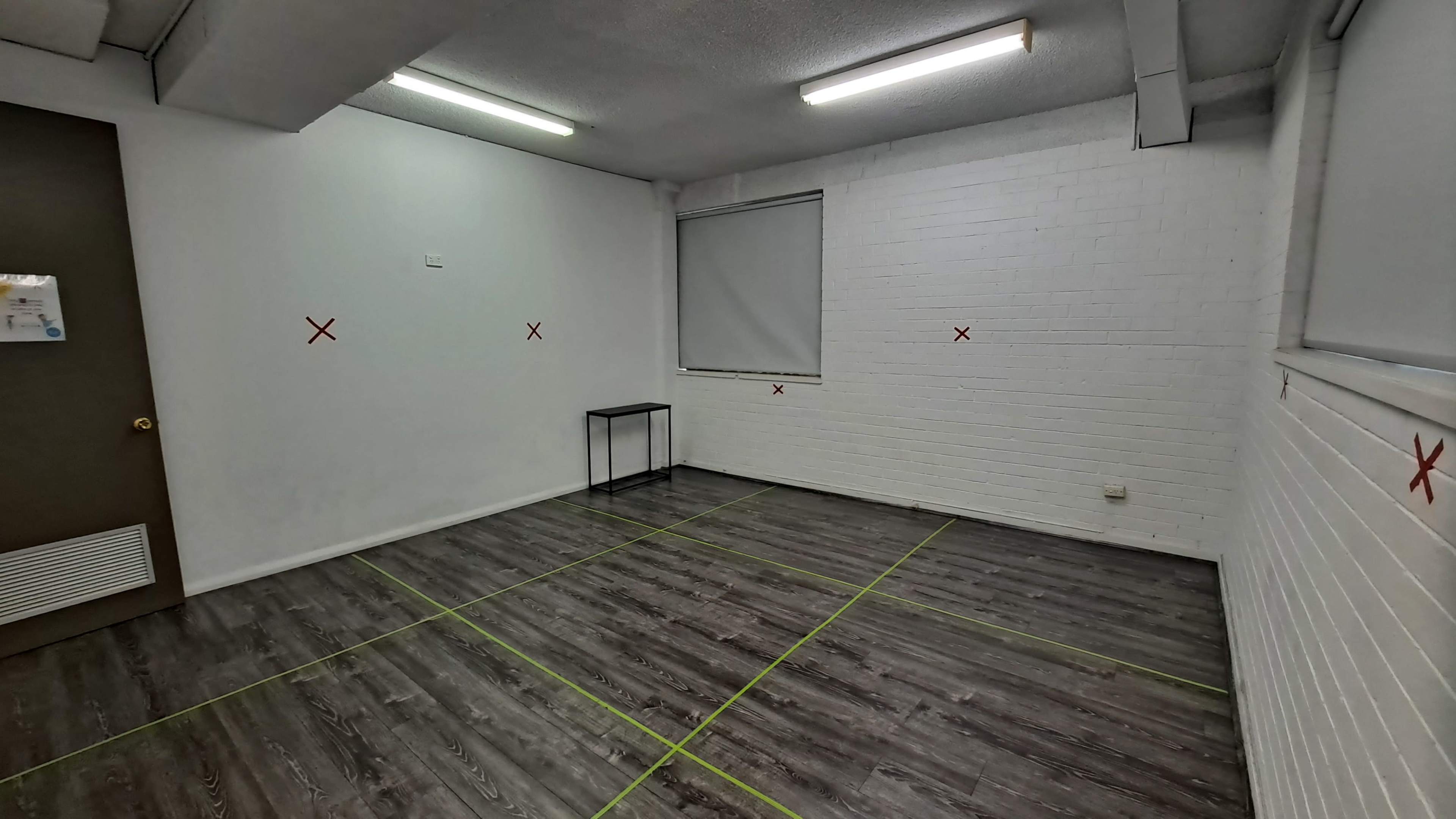 An empty room with white walls, a window covered by a blind, a small black table, and green tape marking boundaries on the floor.