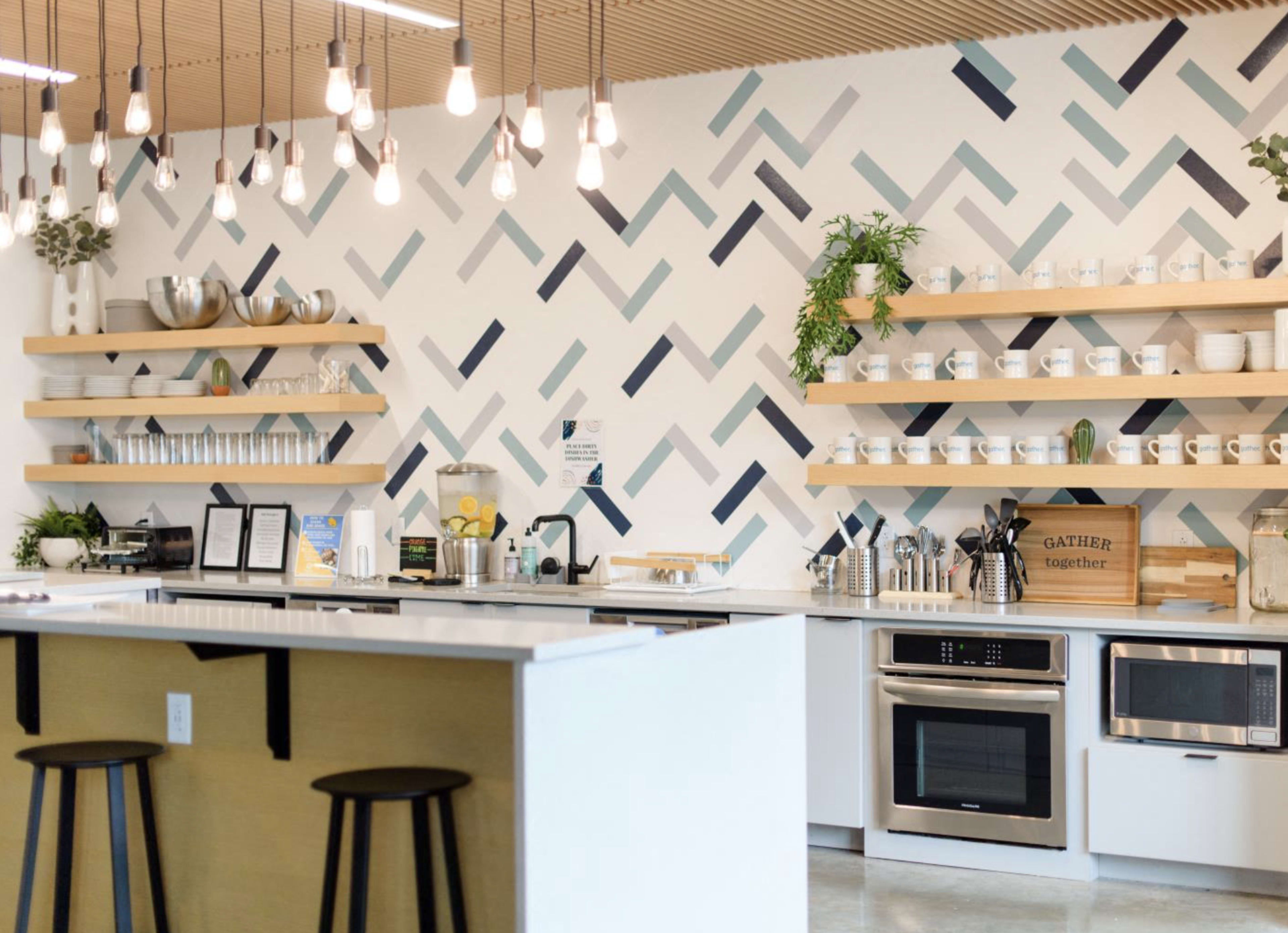 The image shows a modern kitchen with a patterned wall, open shelves displaying dishware, and a central island with bar stools.