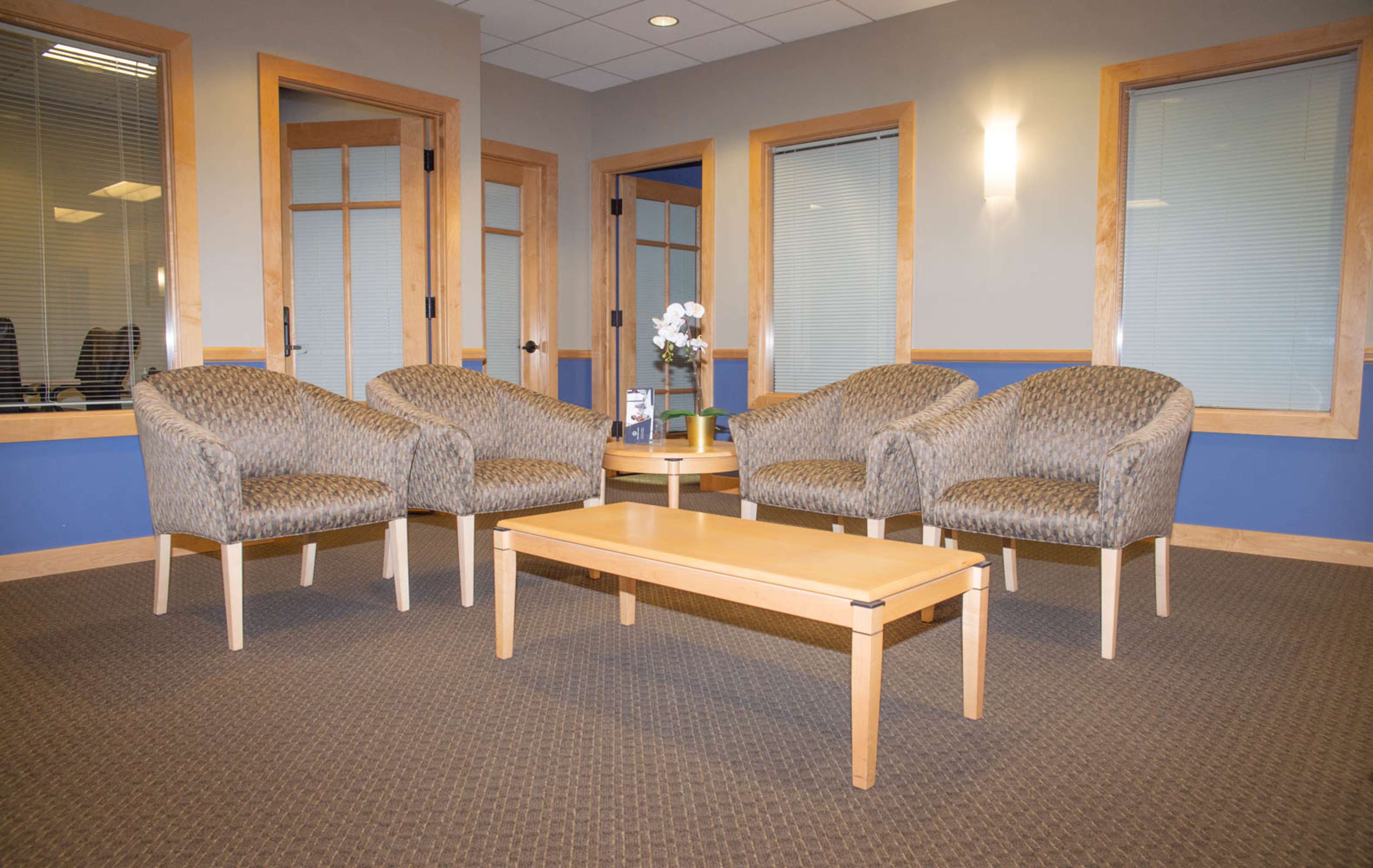 A waiting area features four patterned chairs arranged around a wooden coffee table, set against a backdrop of blue and beige walls.