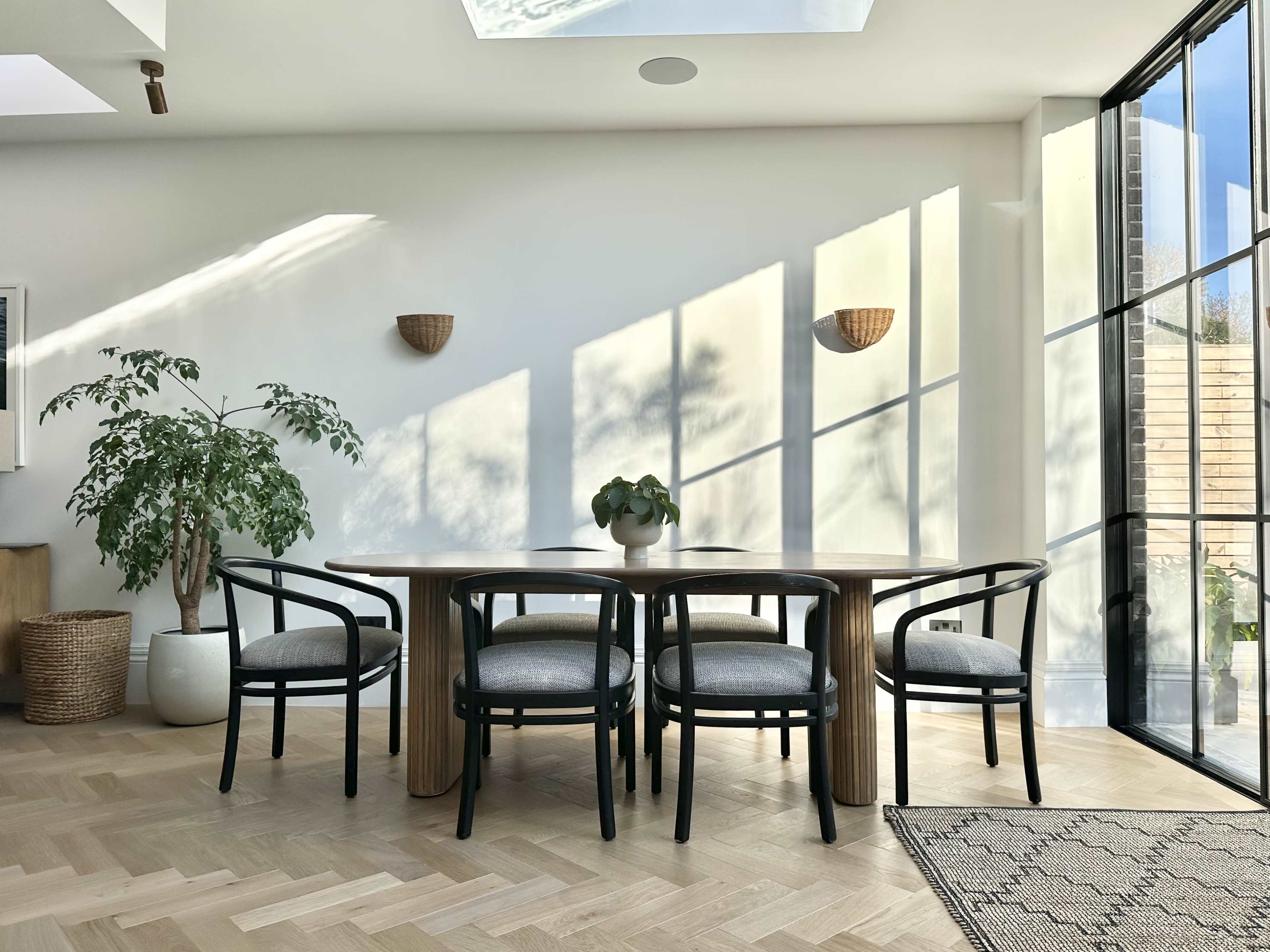 A modern dining area features a round wooden table surrounded by four black chairs, with large windows allowing natural light to cast shadows on the walls.