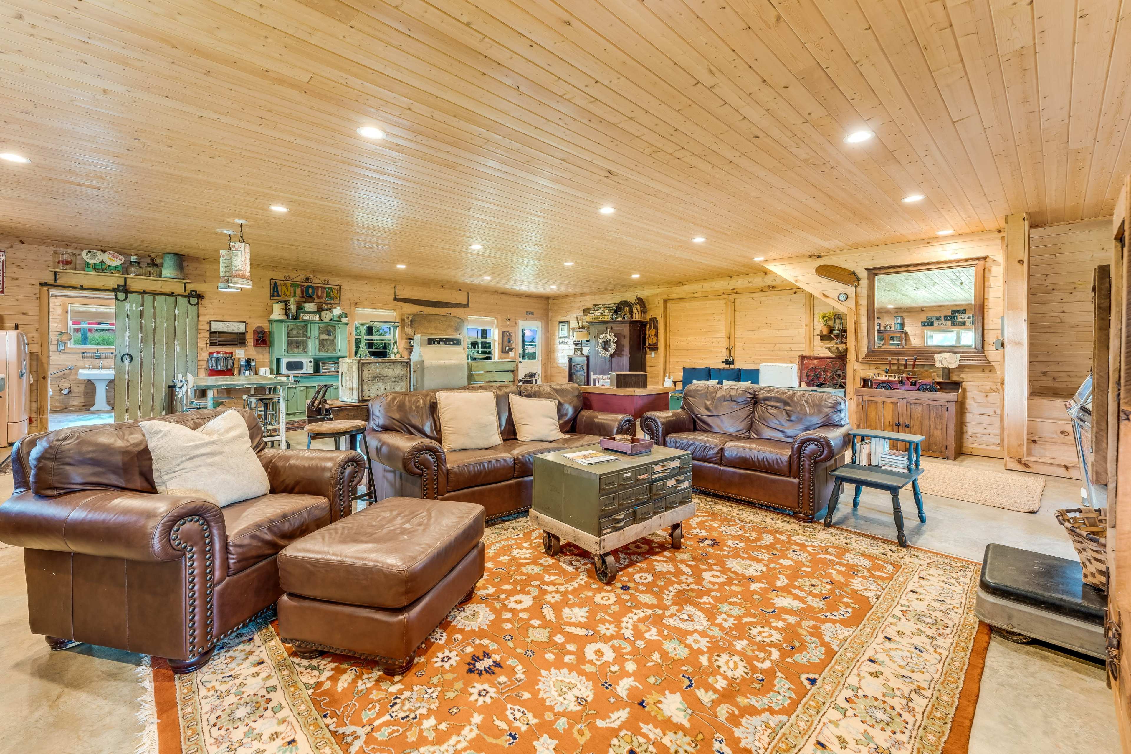 The image shows a large, well-lit living area featuring a brown leather sectional sofa, a patterned area rug, and a rustic wooden table, with a kitchen visible in the background.