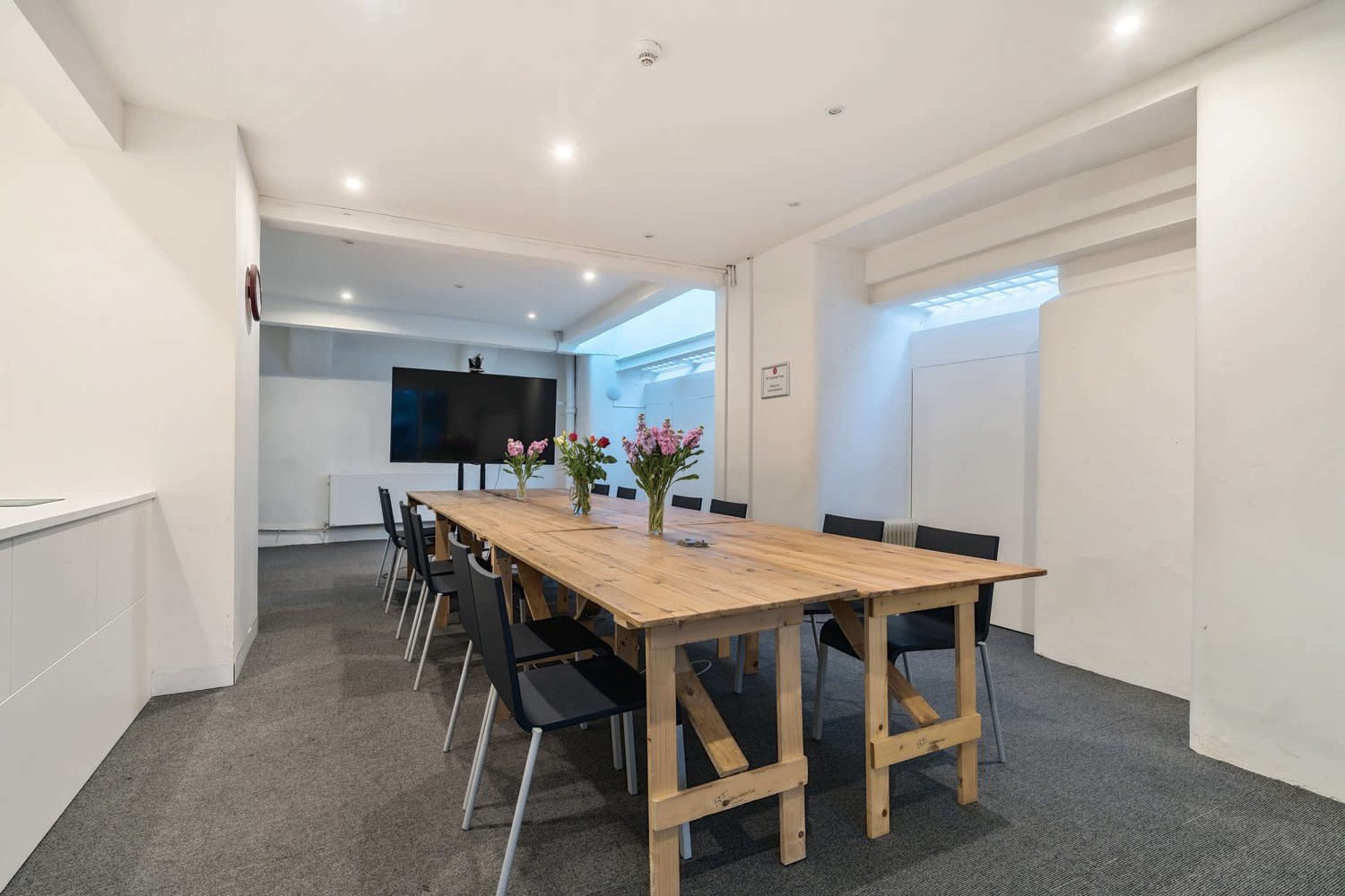The image shows a modern conference room with a large wooden table surrounded by black chairs and a flat-screen television mounted on the wall.