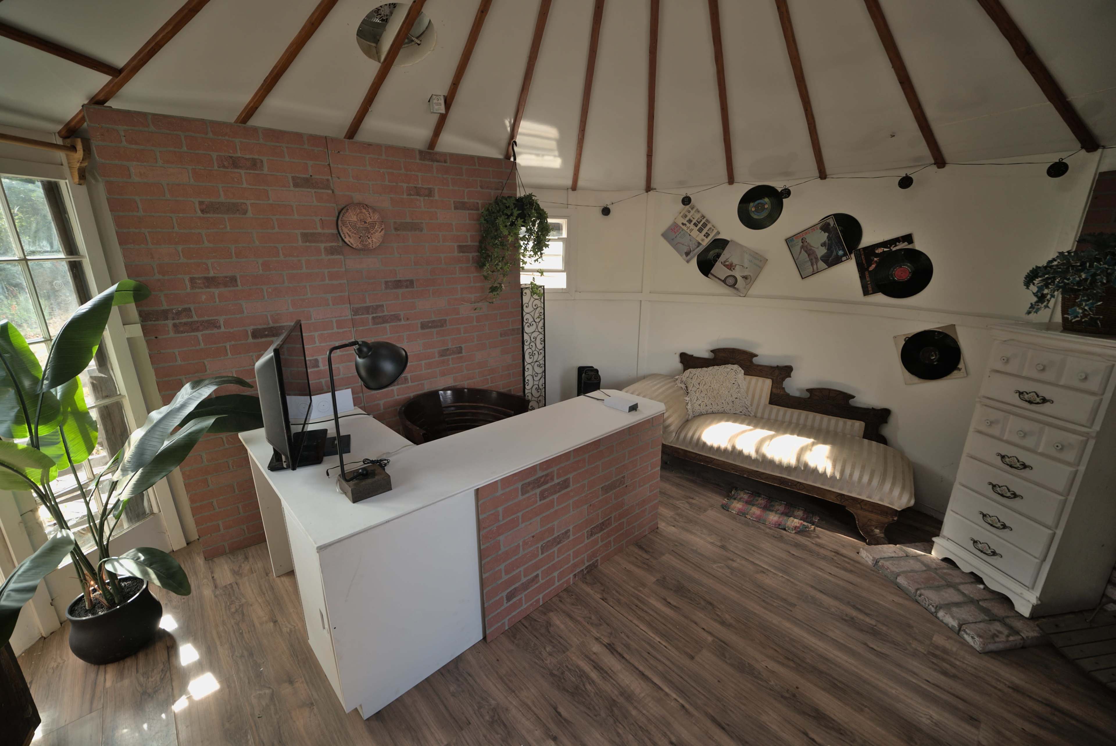 The scene shows a small, circular room with a brick feature wall, a desk with a computer and lamp, a couch, and decorative vinyl records on the walls.