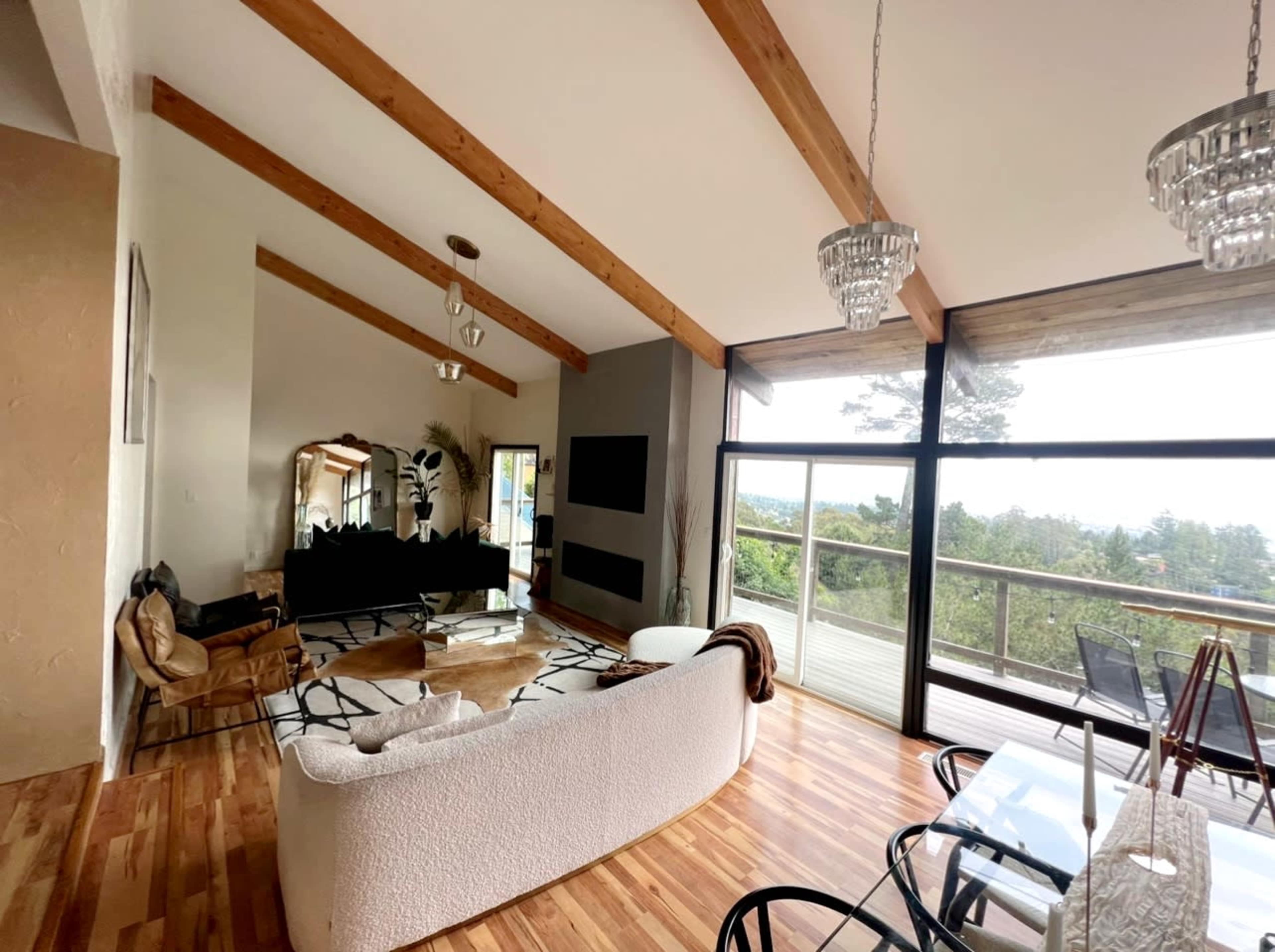 A modern living room with vaulted wooden beams, large windows overlooking greenery, and stylish furniture including a curved white sofa and a glass dining table.