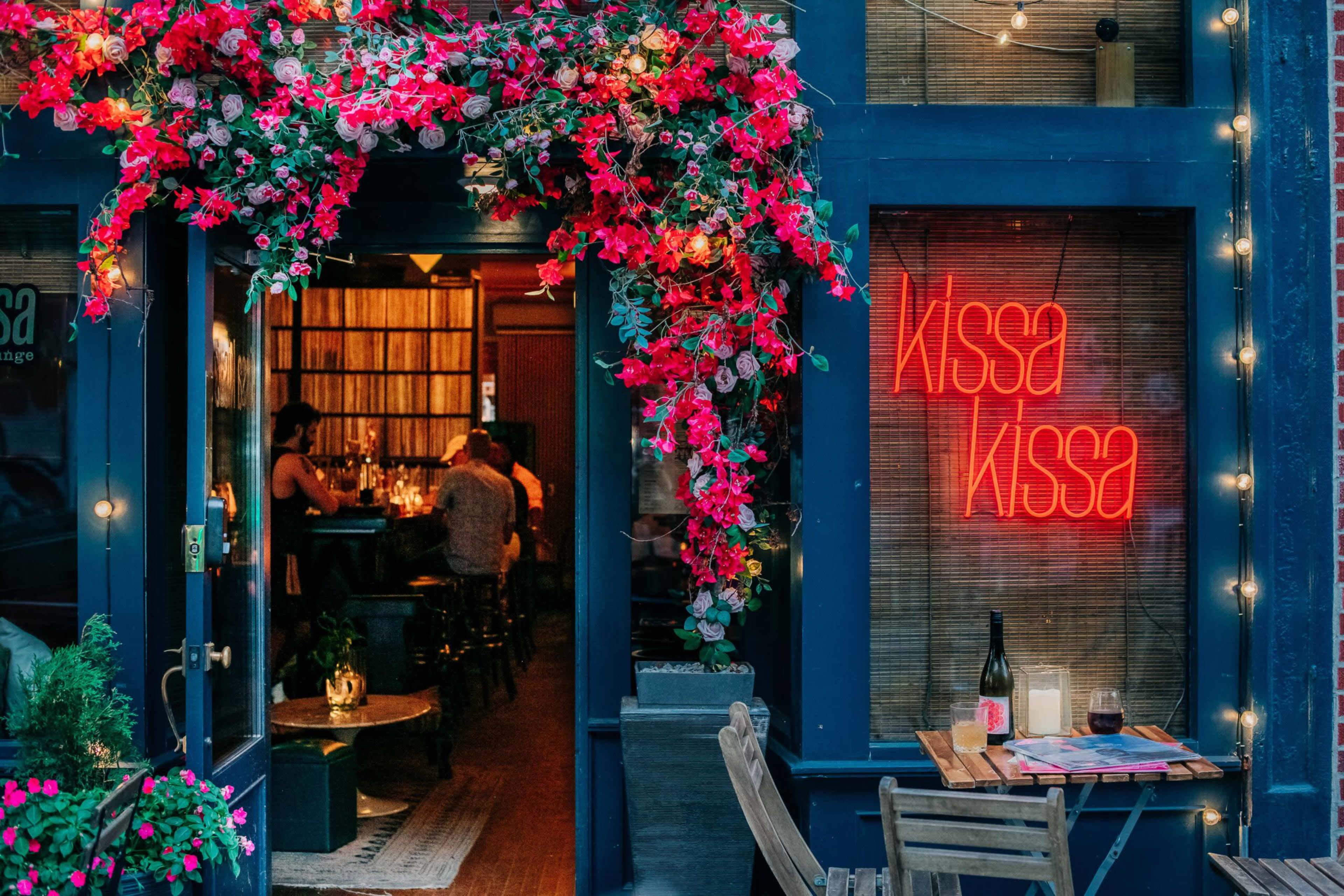 A vibrant entrance of a restaurant features a floral display above the door and a glowing neon sign reading "kissa kissa."