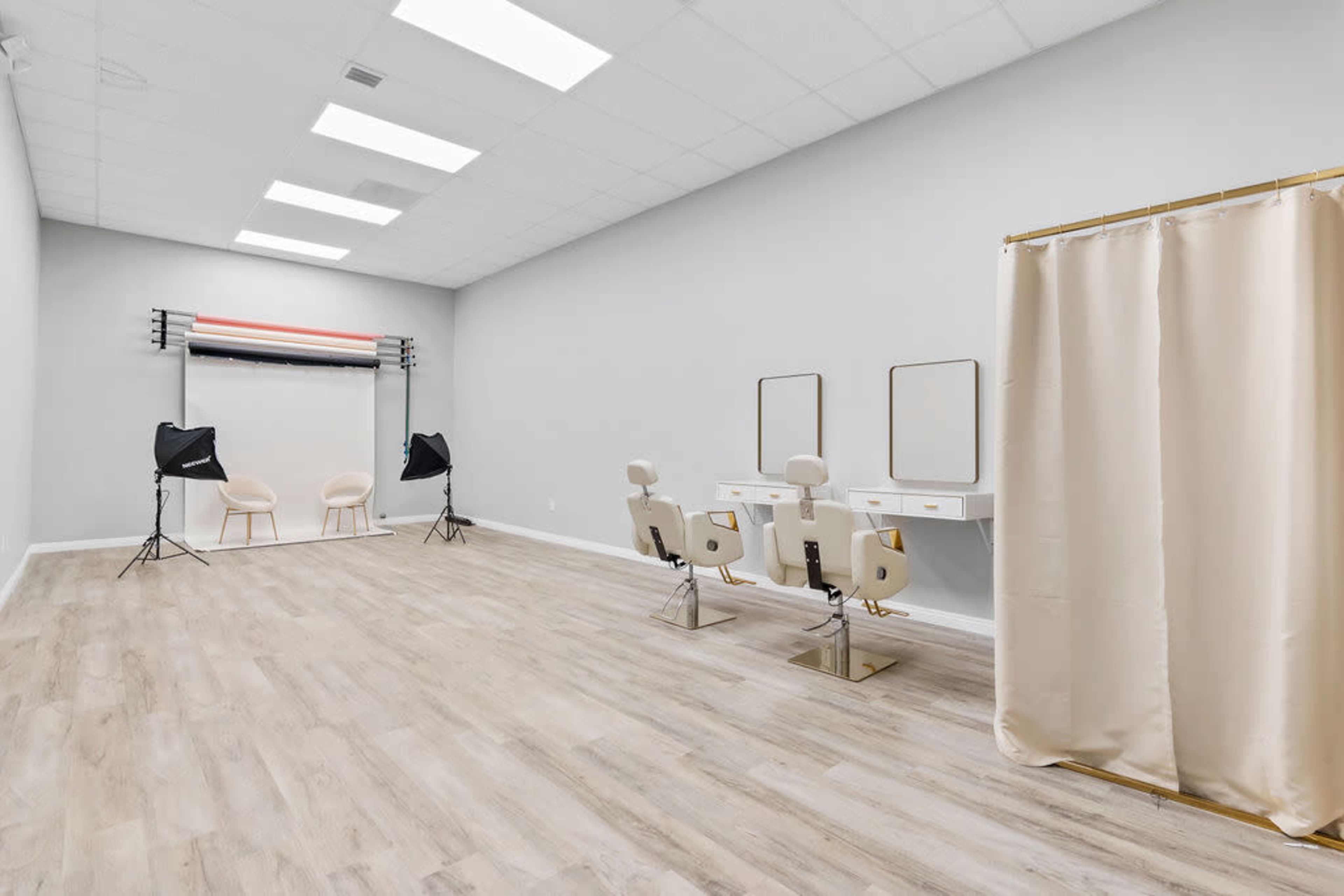 A spacious, minimalist studio interior with two hair styling stations, a backdrop setup, and two photography lights.