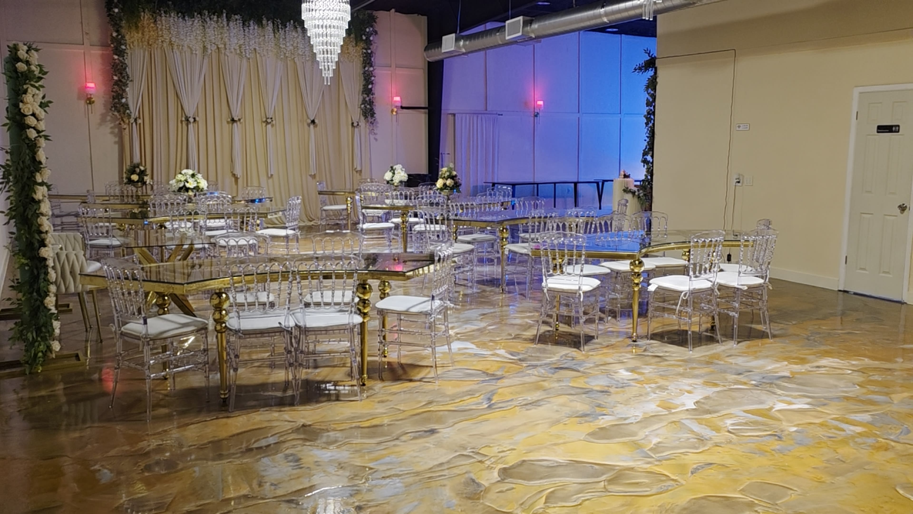 The image shows a spacious event hall with gold and clear acrylic tables and chairs, illuminated by a chandelier and soft lighting, featuring a polished floor.