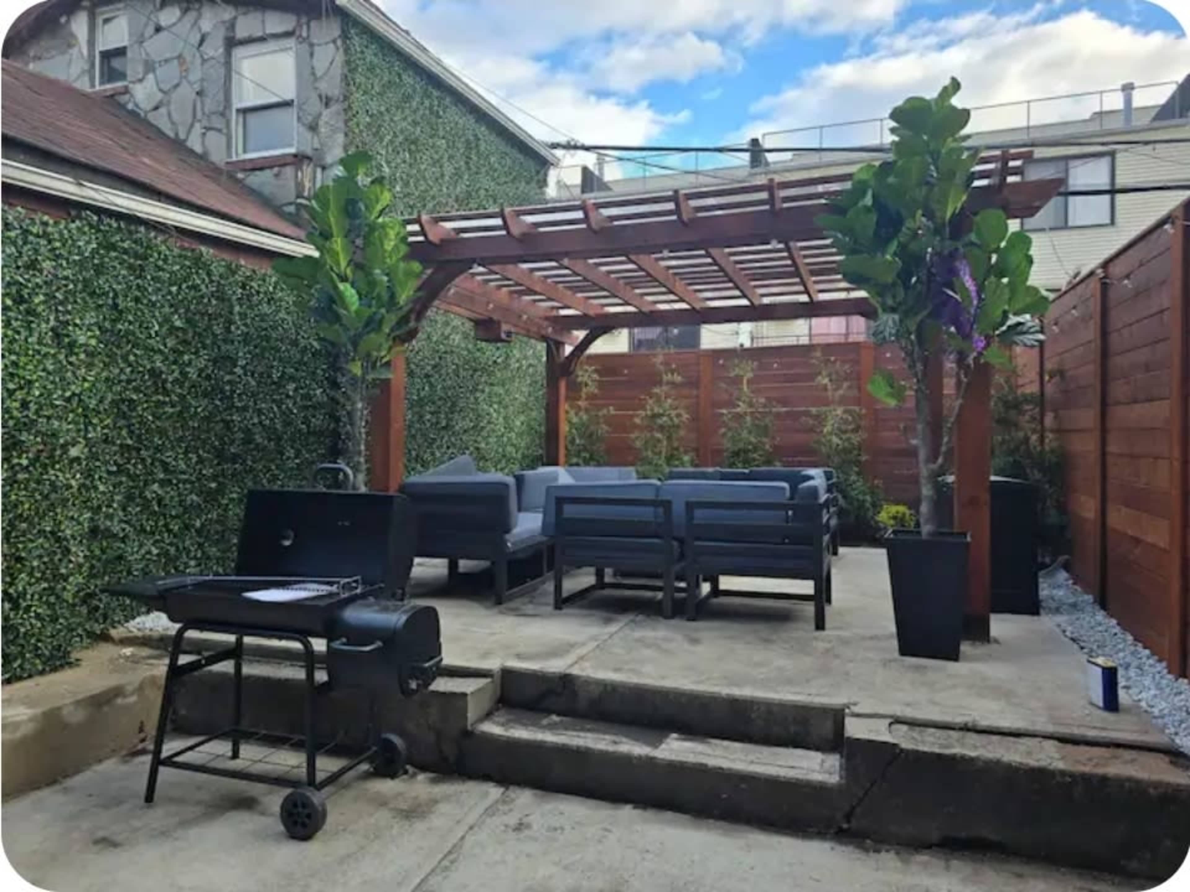 The image shows a patio area with a wooden pergola, lounge seating, a grill, and greenery along the walls.