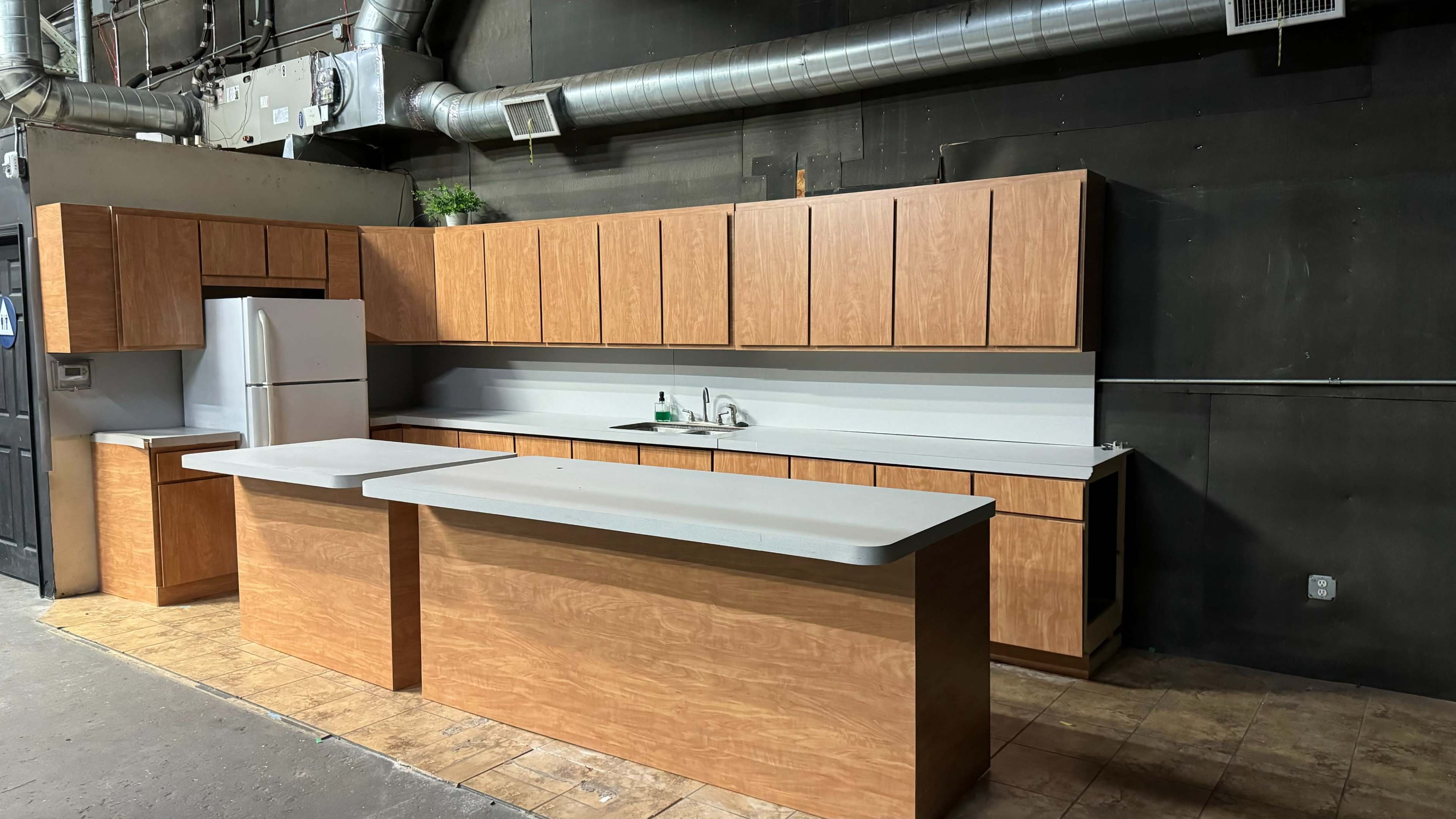 The image shows a simple kitchen setup with wooden cabinets, a countertop island, and basic appliances, including a refrigerator and sink.