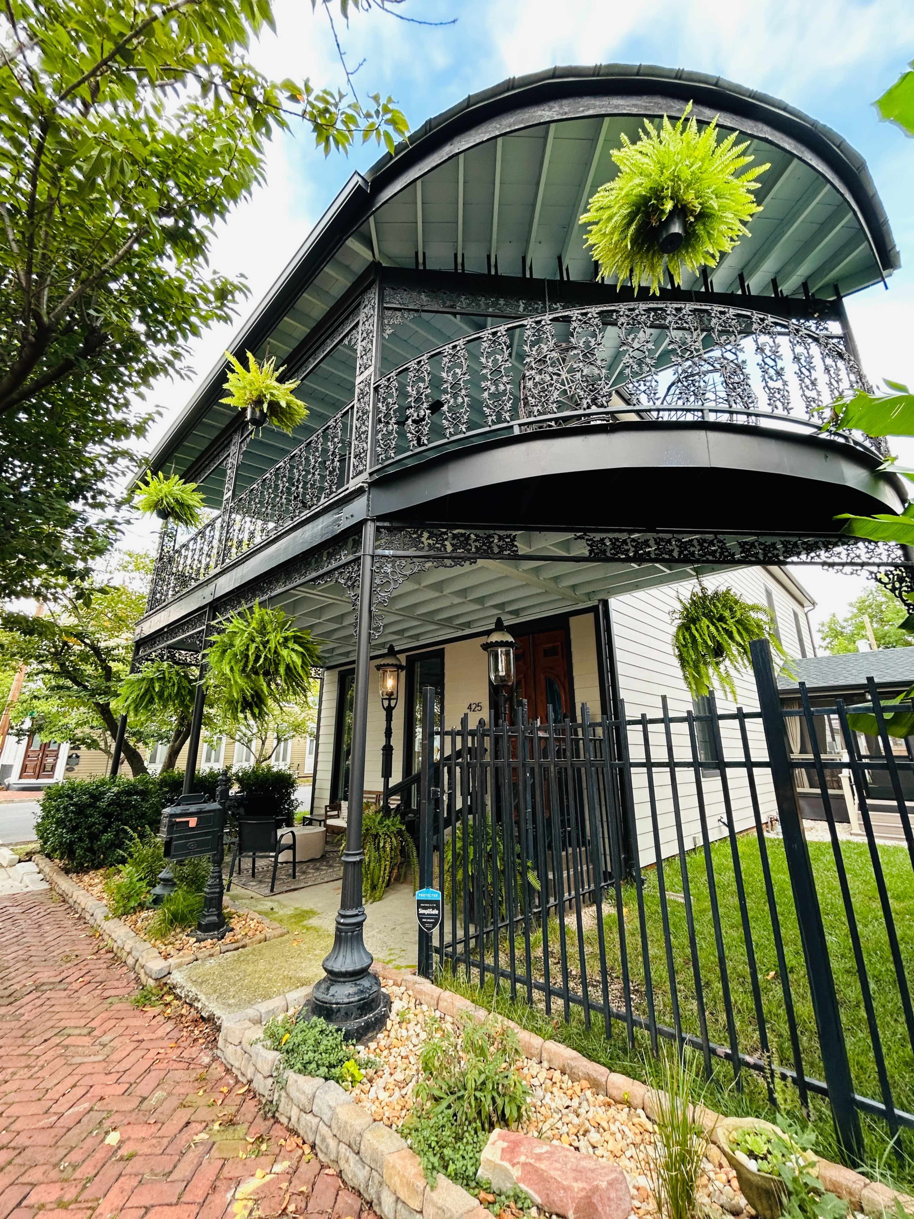 The image shows a two-story house with an ornate wrought iron balcony and hanging ferns, surrounded by a decorative fence and a brick walkway.