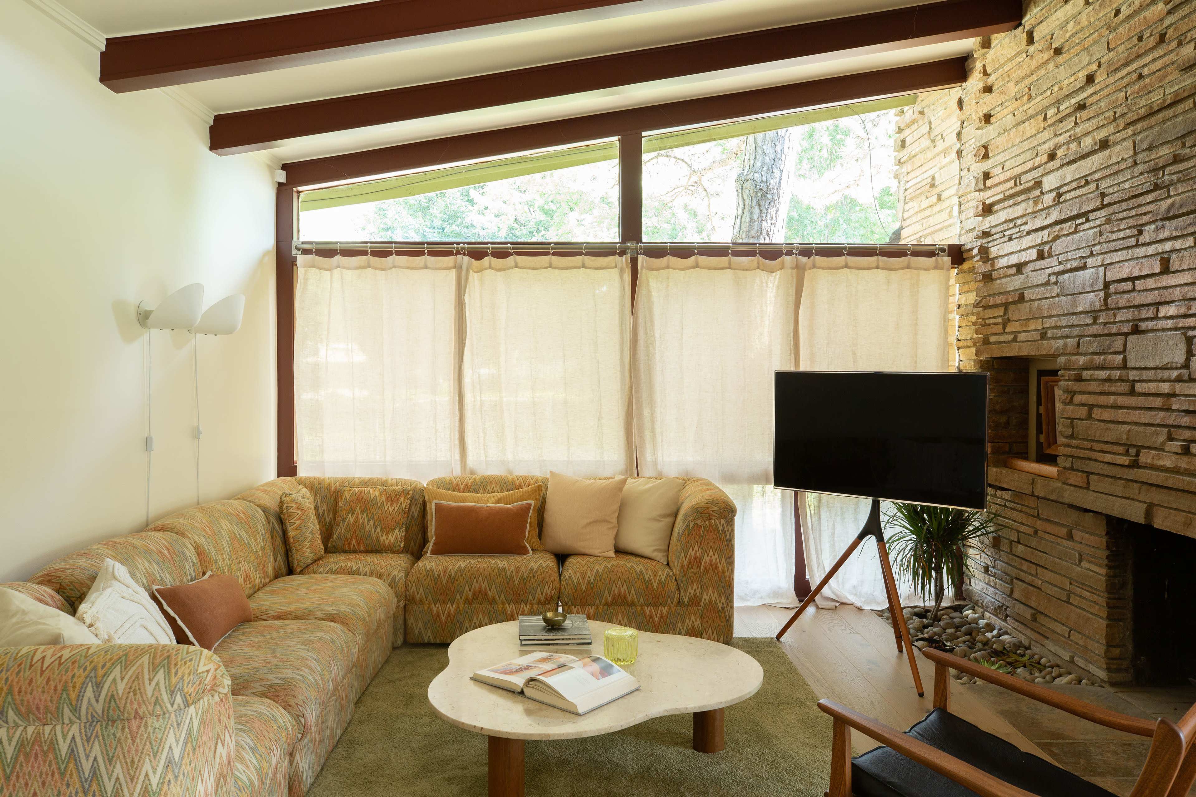 The image shows a living room with a large sectional sofa, a coffee table, a television on a stand, and large windows covered with light curtains.