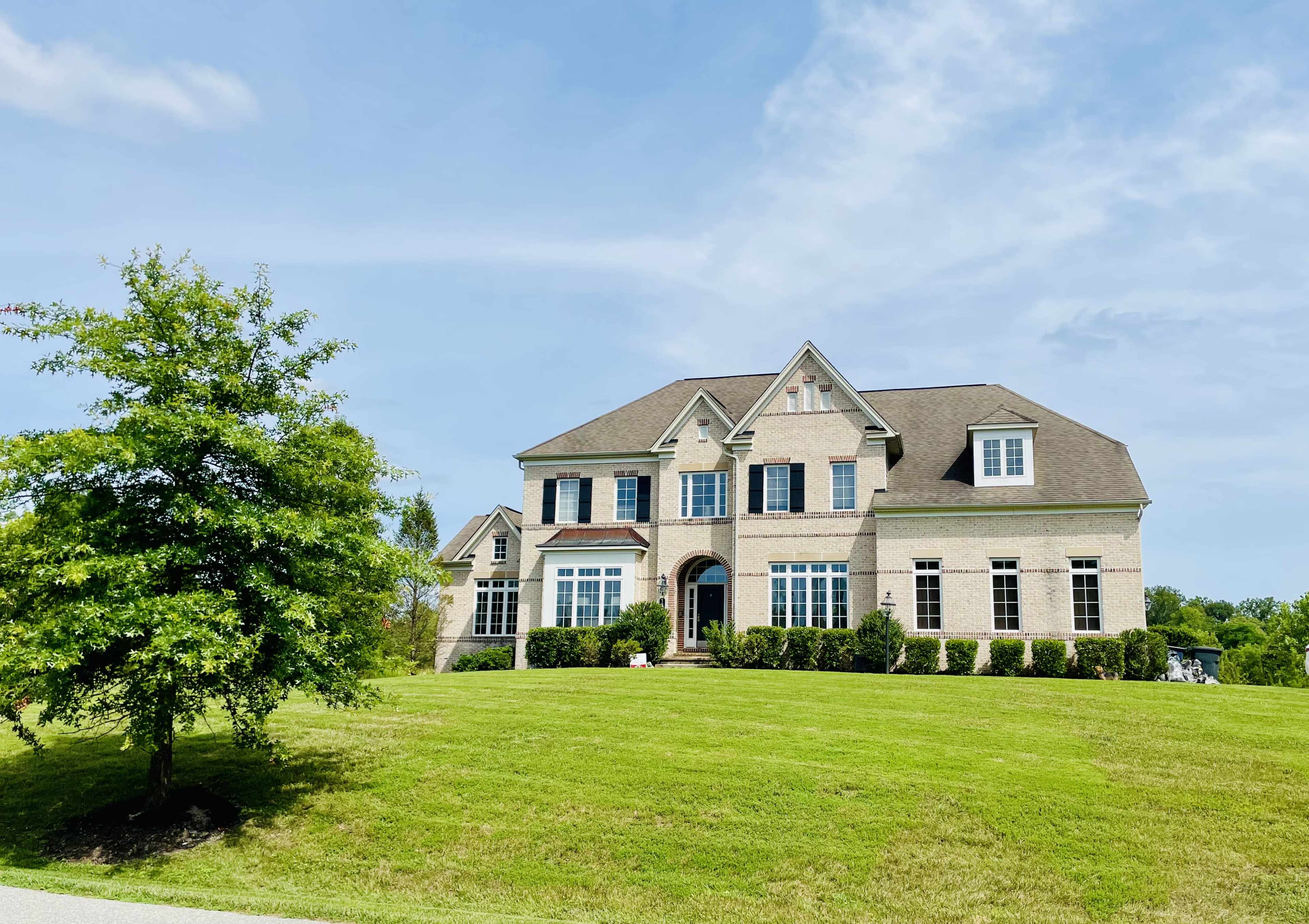 A large brick house with multiple windows is situated on a grassy hill, surrounded by neatly trimmed hedges and a tree in the foreground.
