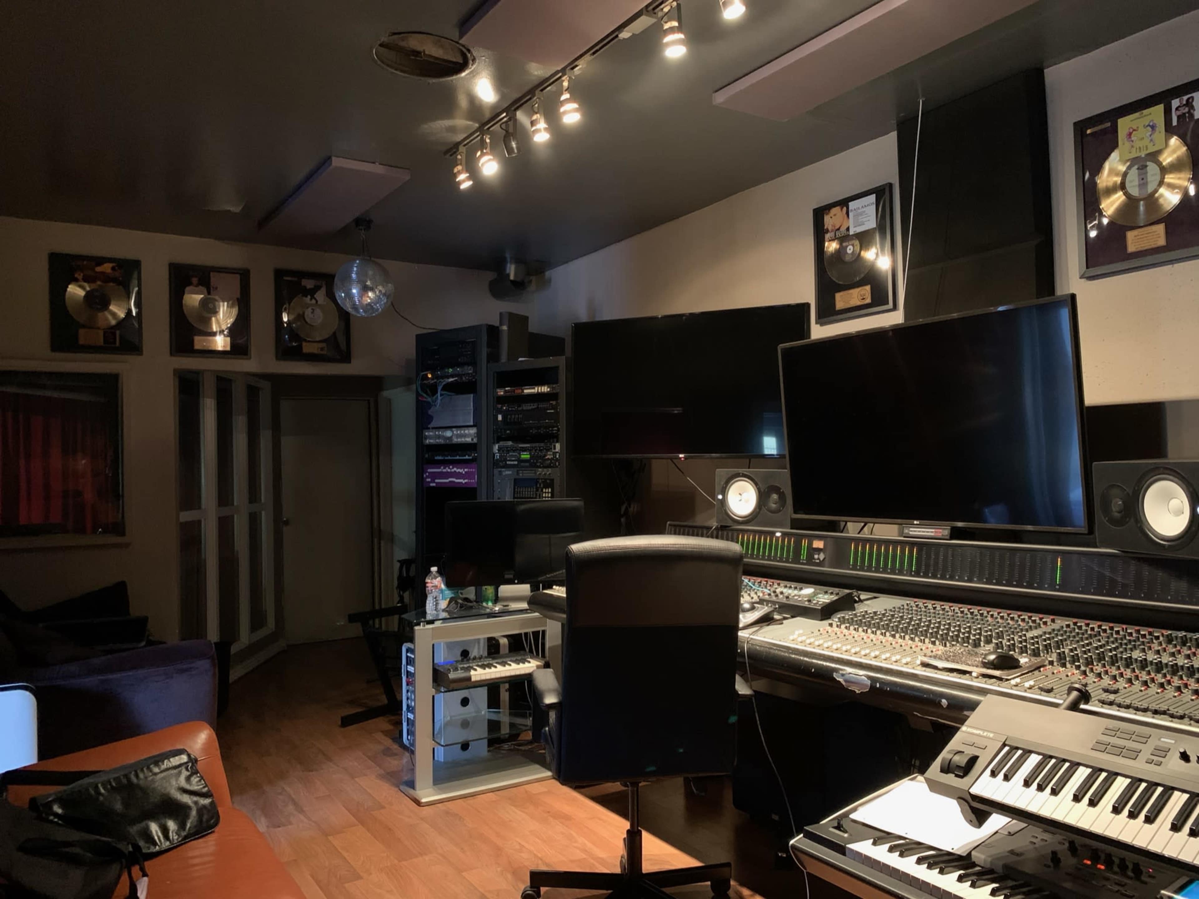 The image shows a music recording studio with a large mixing console, dual monitors, and several gold record plaques on the walls.