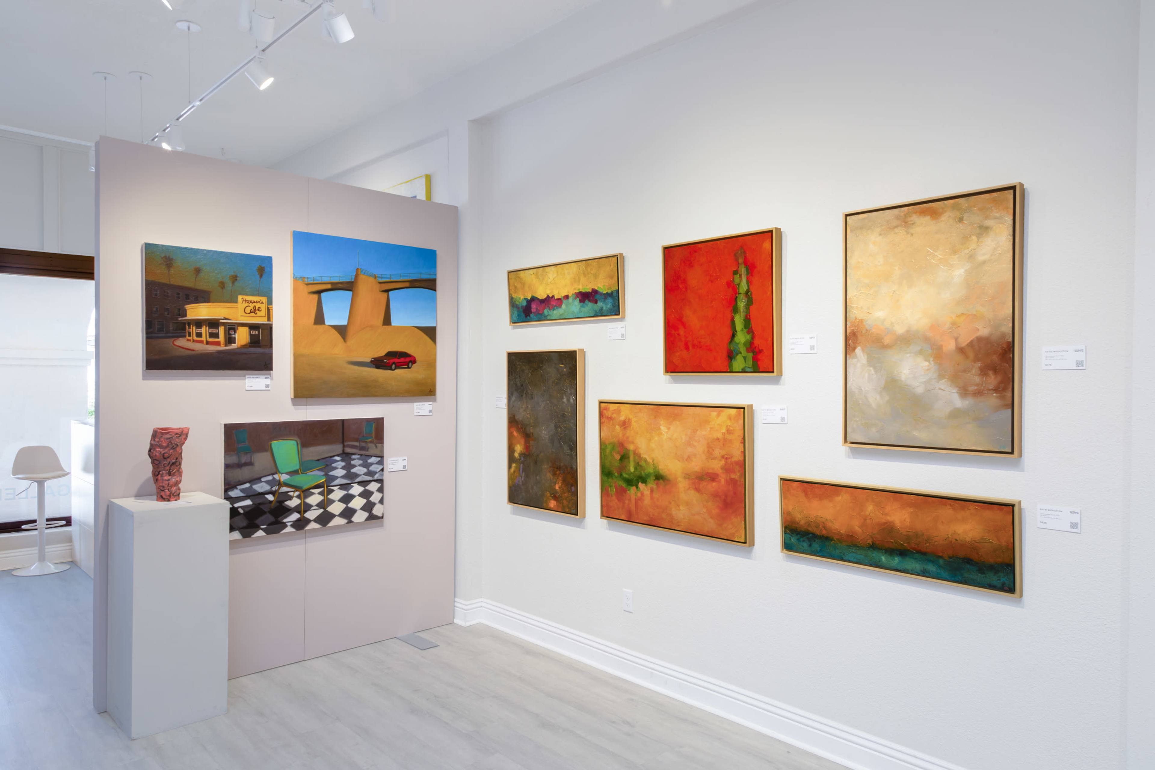 The image depicts an art gallery featuring colorful abstract paintings displayed on white walls, with sculptures and a large photograph accentuating the space.