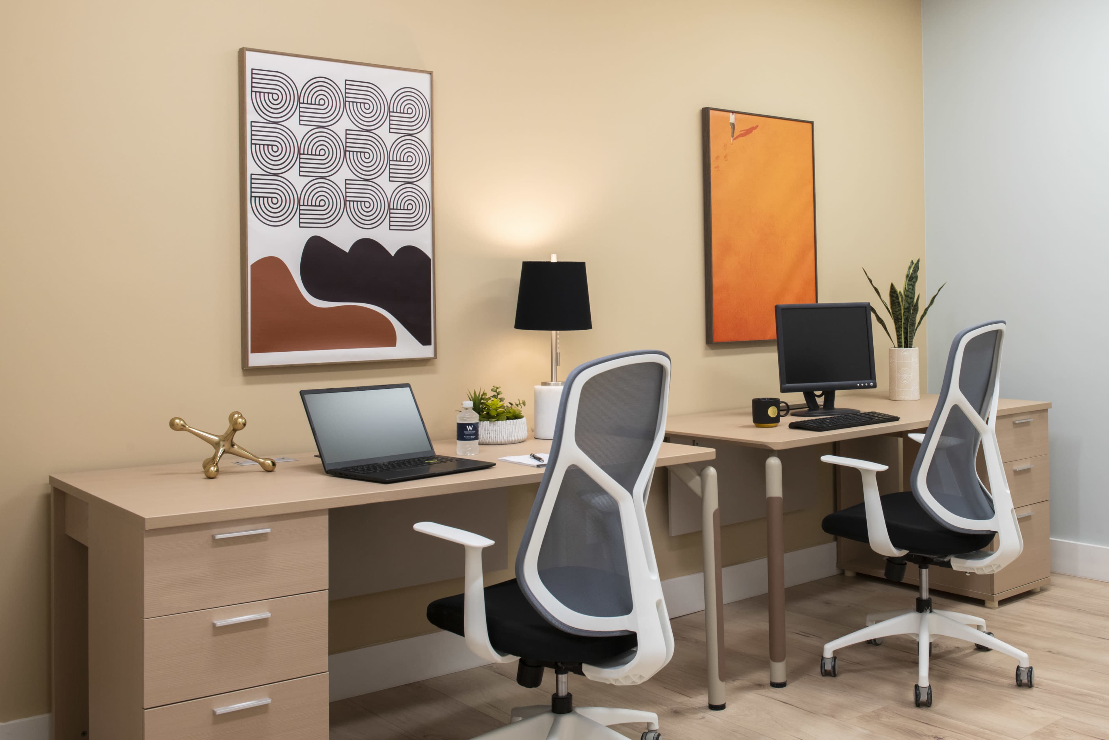 The image depicts a minimalist office space featuring two desks, each with a chair, a laptop, a computer monitor, and decorative art on the walls.