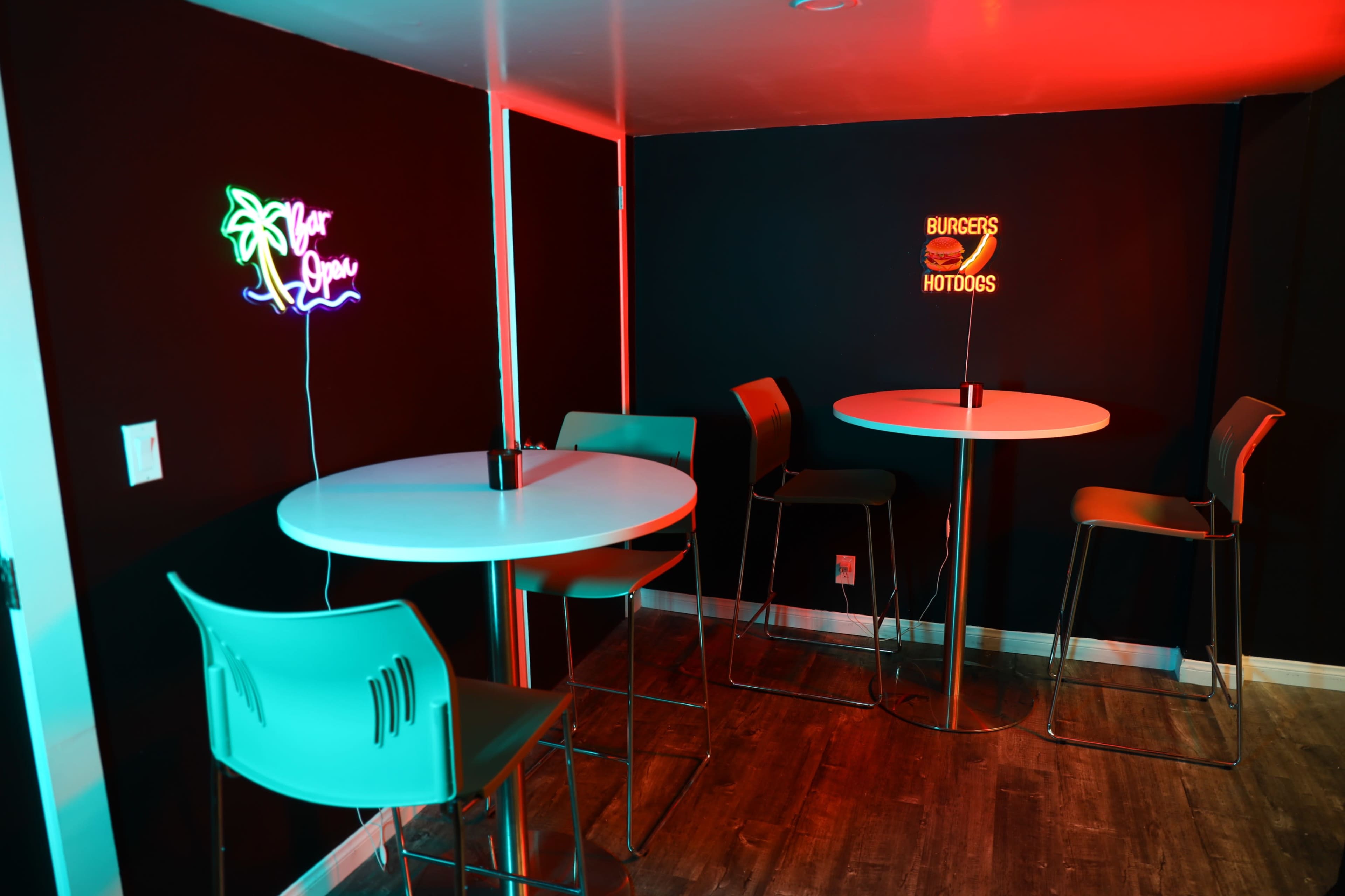 The image shows a small seating area with tall tables and chairs, illuminated by neon signs that read "Open" and "Burgers & Hotdogs."