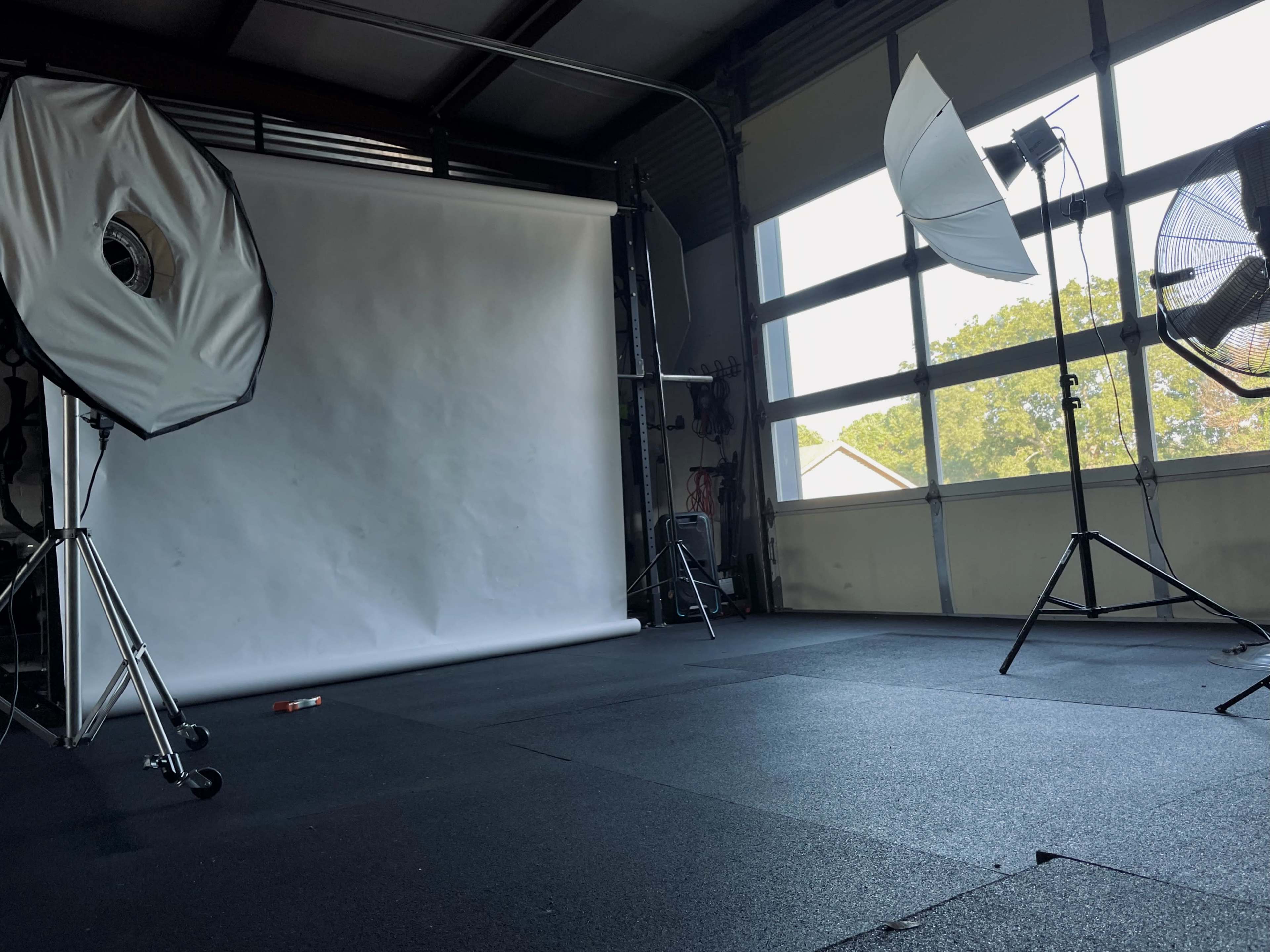 The image shows a photography studio with two softbox lights, a backdrop stand with a white paper roll, and two large fans positioned near a window.