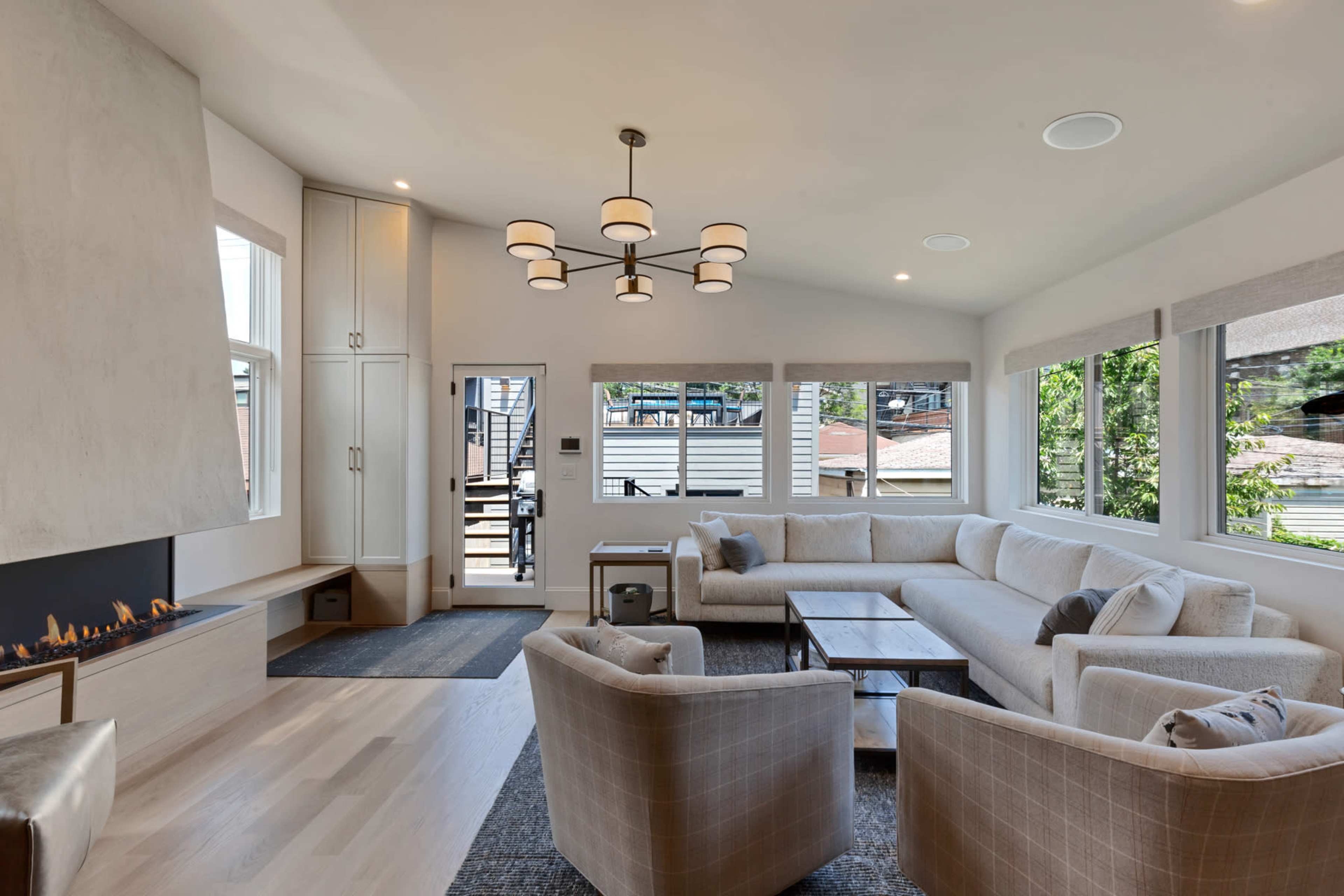 The image shows a modern living room with a large sectional sofa, a coffee table, a contemporary light fixture, and large windows that overlook an outdoor area.