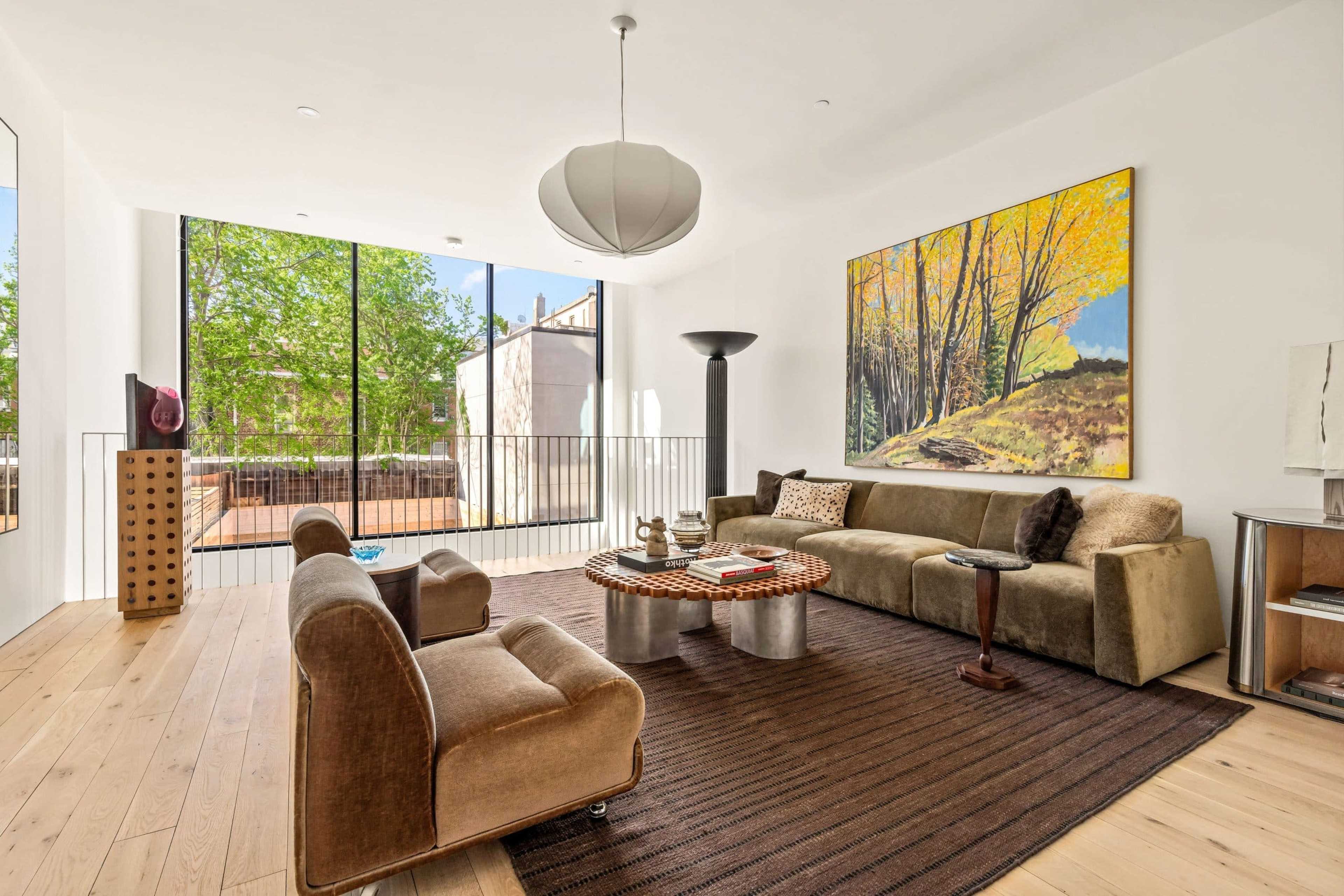 A modern living room features a large window with views of greenery, a sectional sofa, two armchairs, a coffee table, and a vibrant landscape painting on the wall.