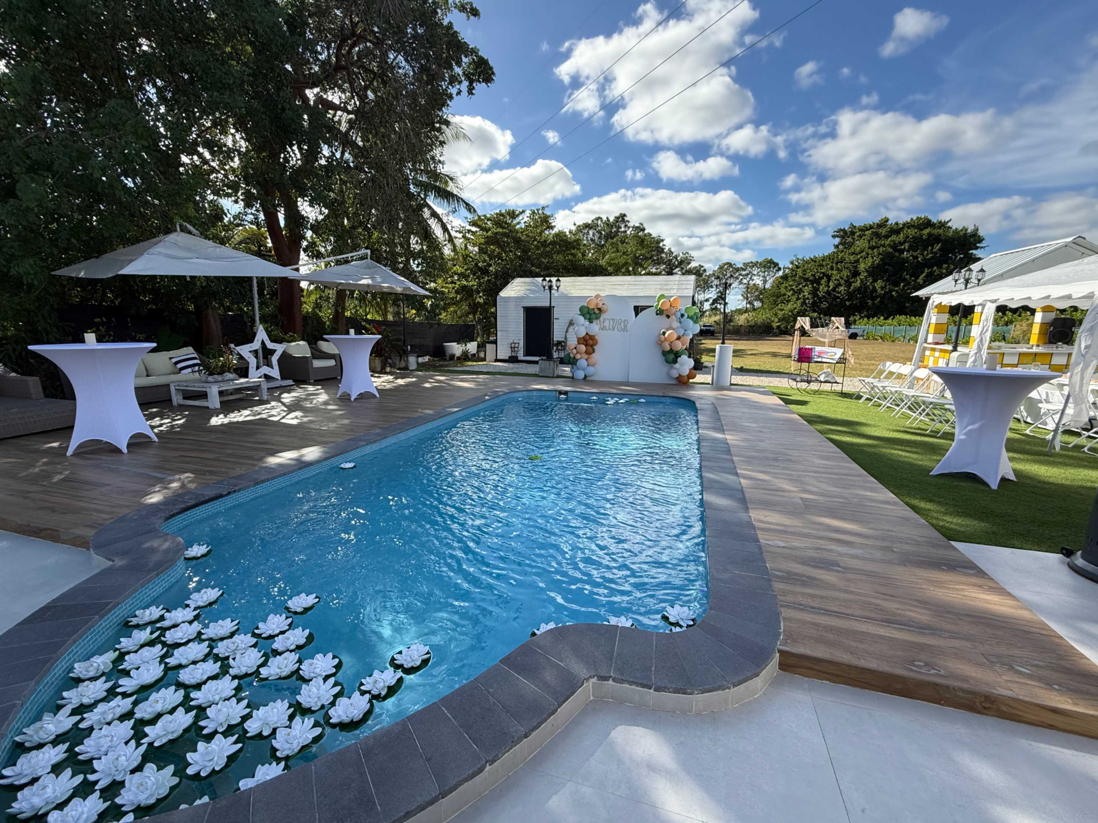 The image shows a well-maintained backyard with a swimming pool surrounded by lounge chairs, white tables, and decorative elements, under a blue sky with scattered clouds.