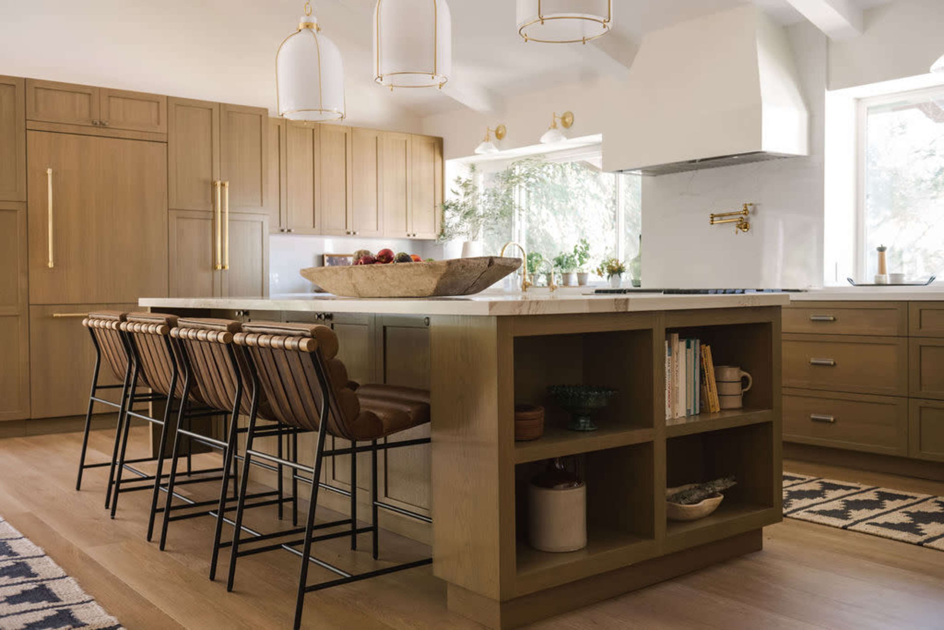 A modern kitchen features a large central island with bar stools, wooden cabinetry, and a white countertop adorned with a bowl of fruit.