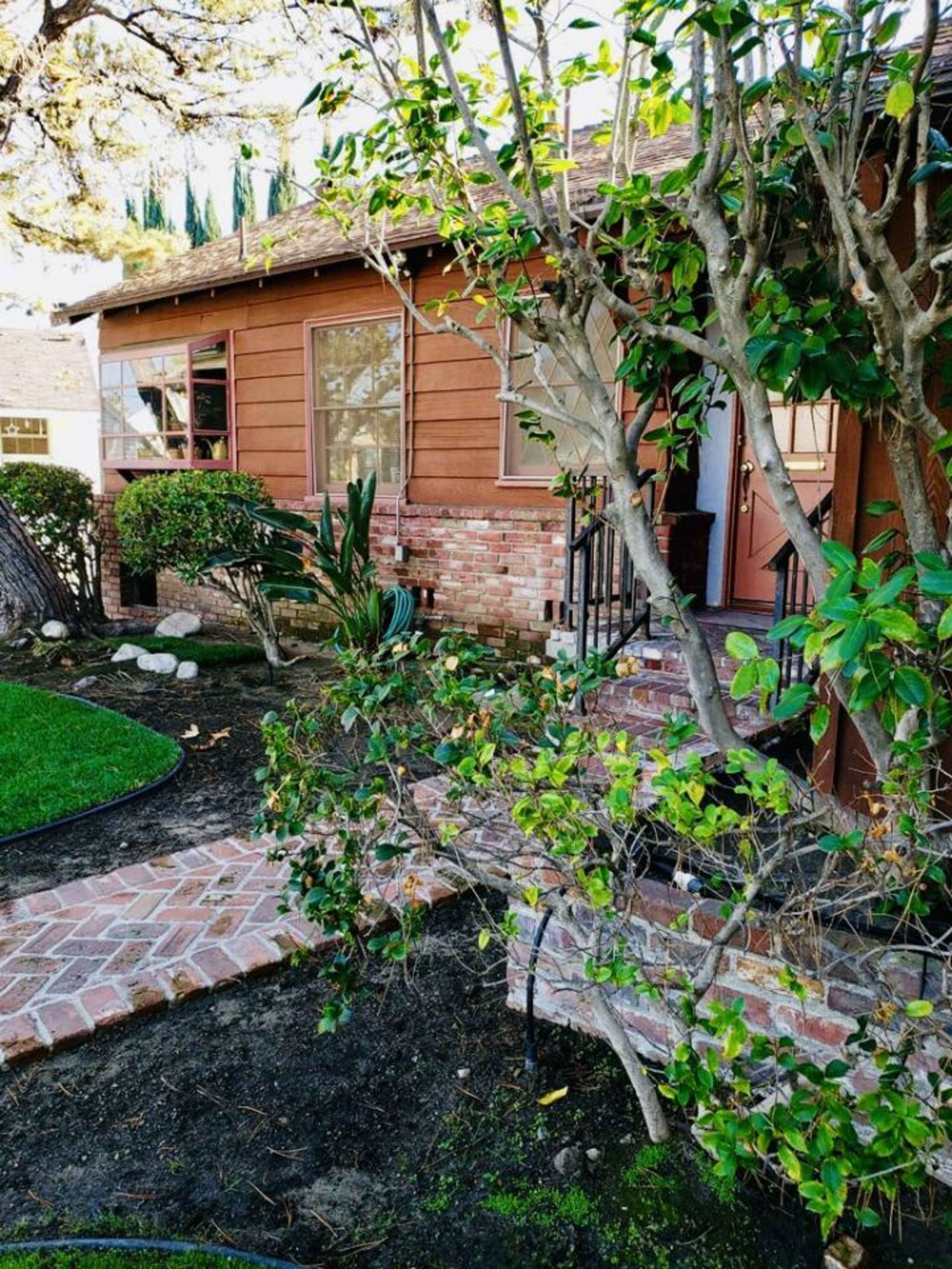 A brown house with brick accents features a walkway bordered by shrubs and small plants leading to its entrance.