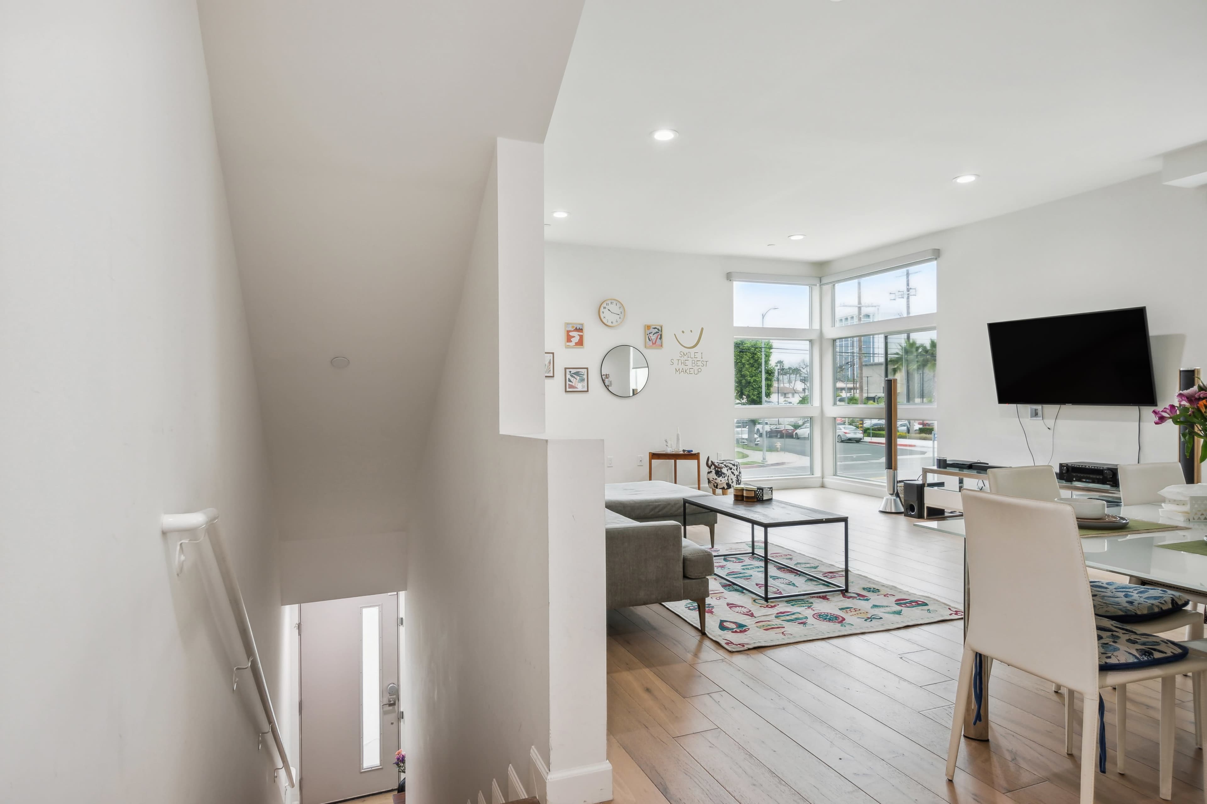 The image shows a modern living area with a staircase leading down, large windows, and a dining table set for meals.