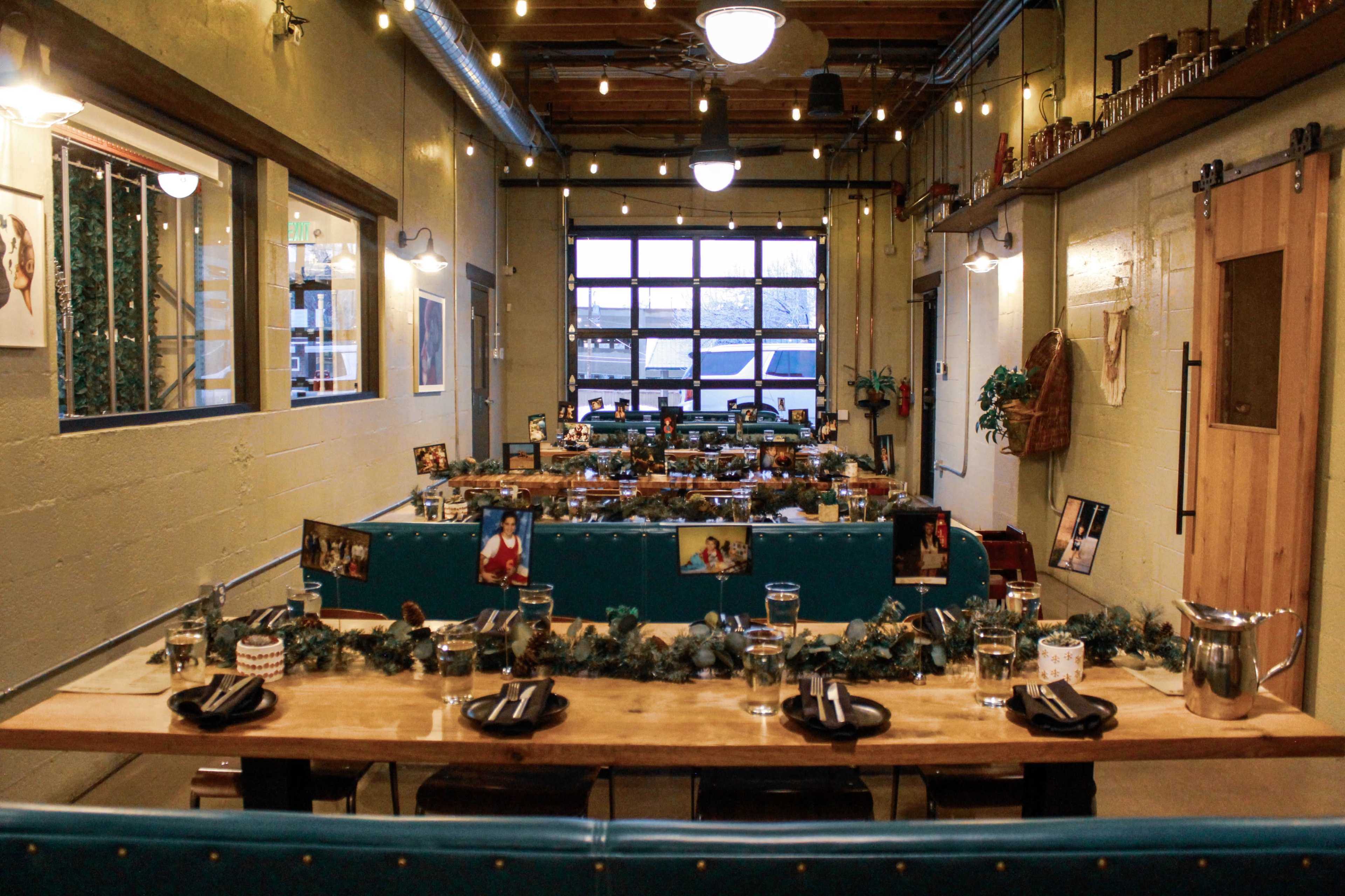 A decorated dining space with wooden tables set for a meal, string lights overhead, and a large garage door at the end of the room.