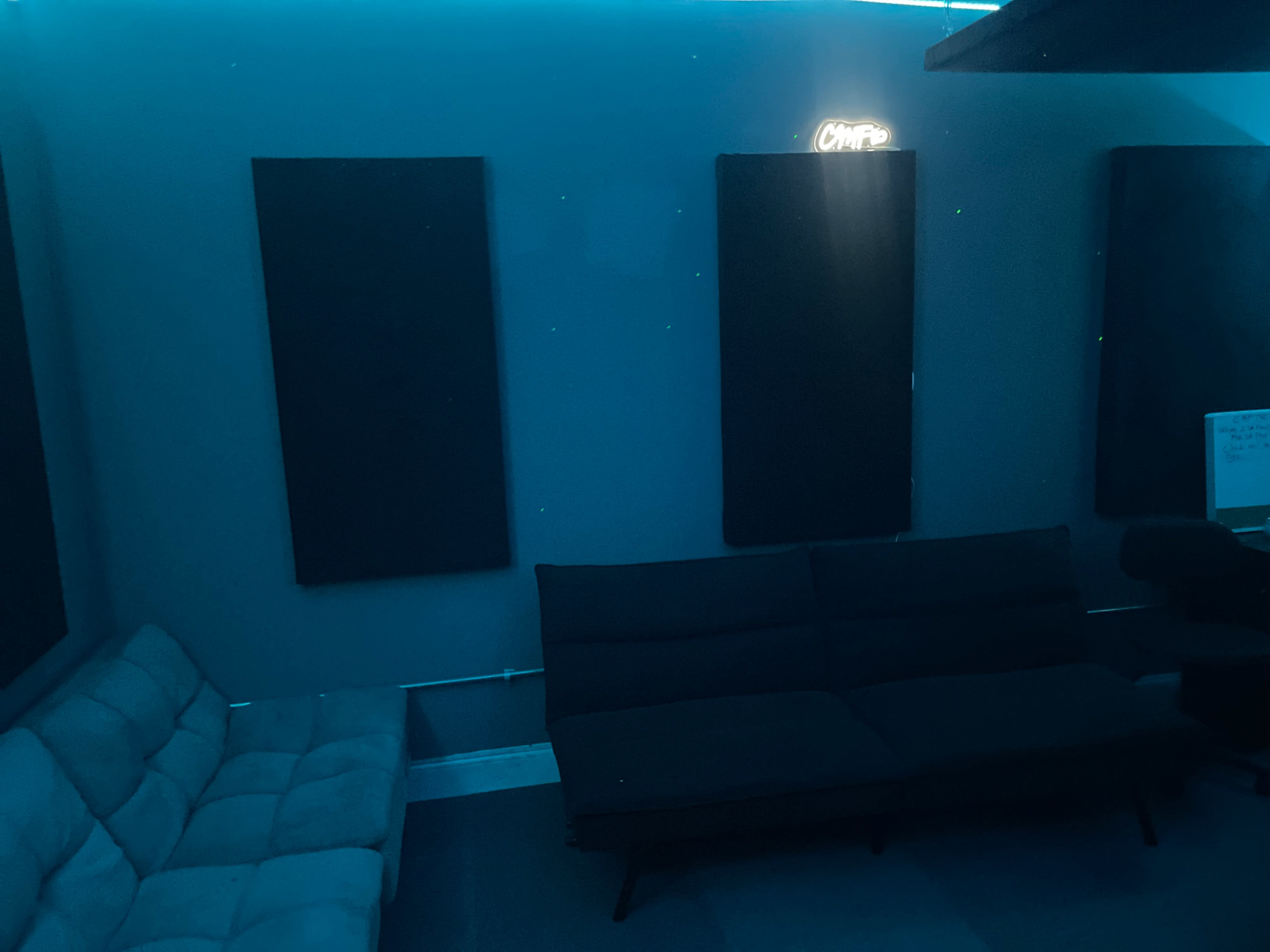 The image shows a dimly lit room with two dark soundproofing panels on the walls, a gray couch, and a black sofa, illuminated by a blue light.