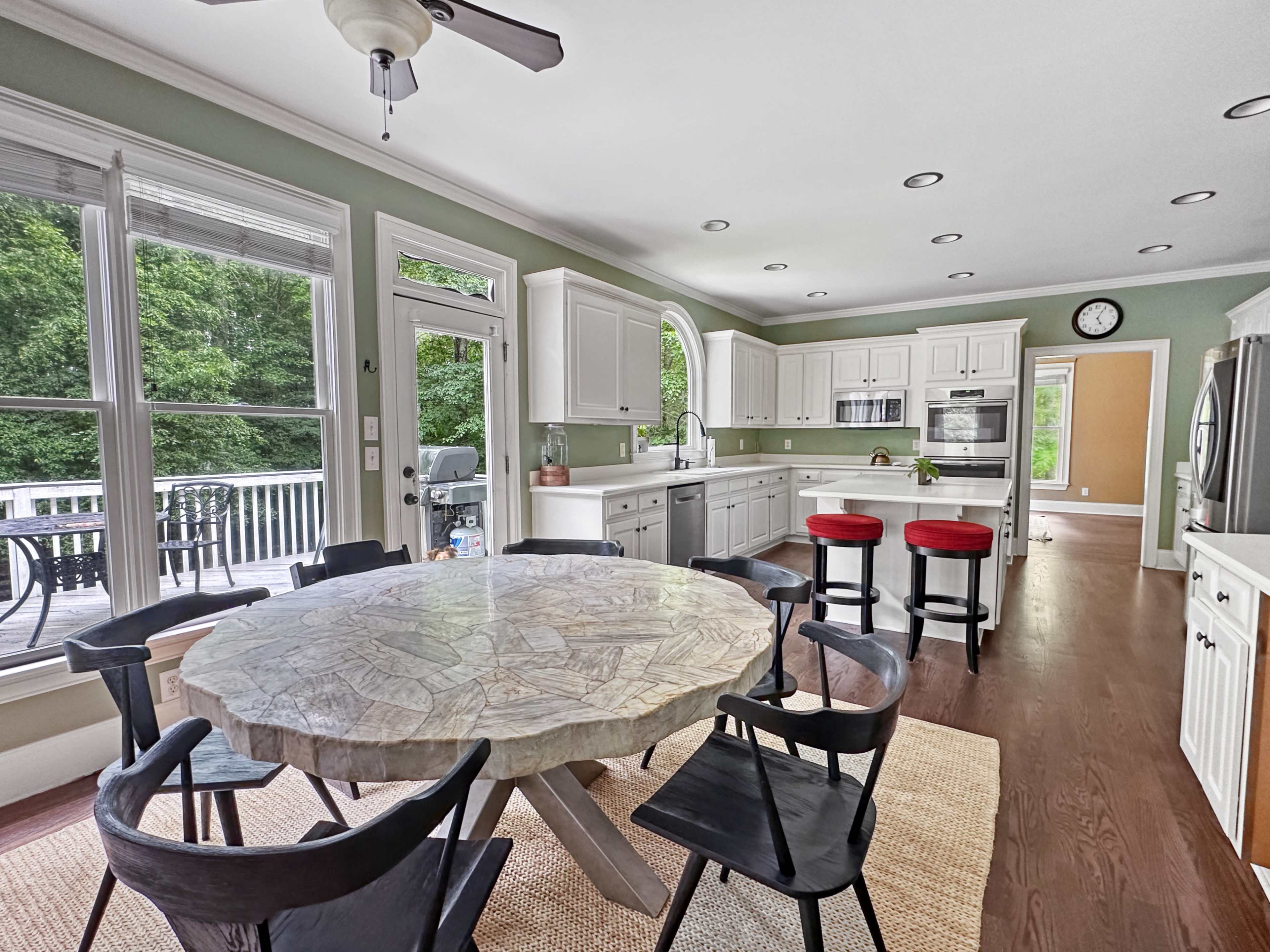 A modern kitchen features a round wooden table surrounded by black chairs, with white cabinetry and large windows overlooking a deck.