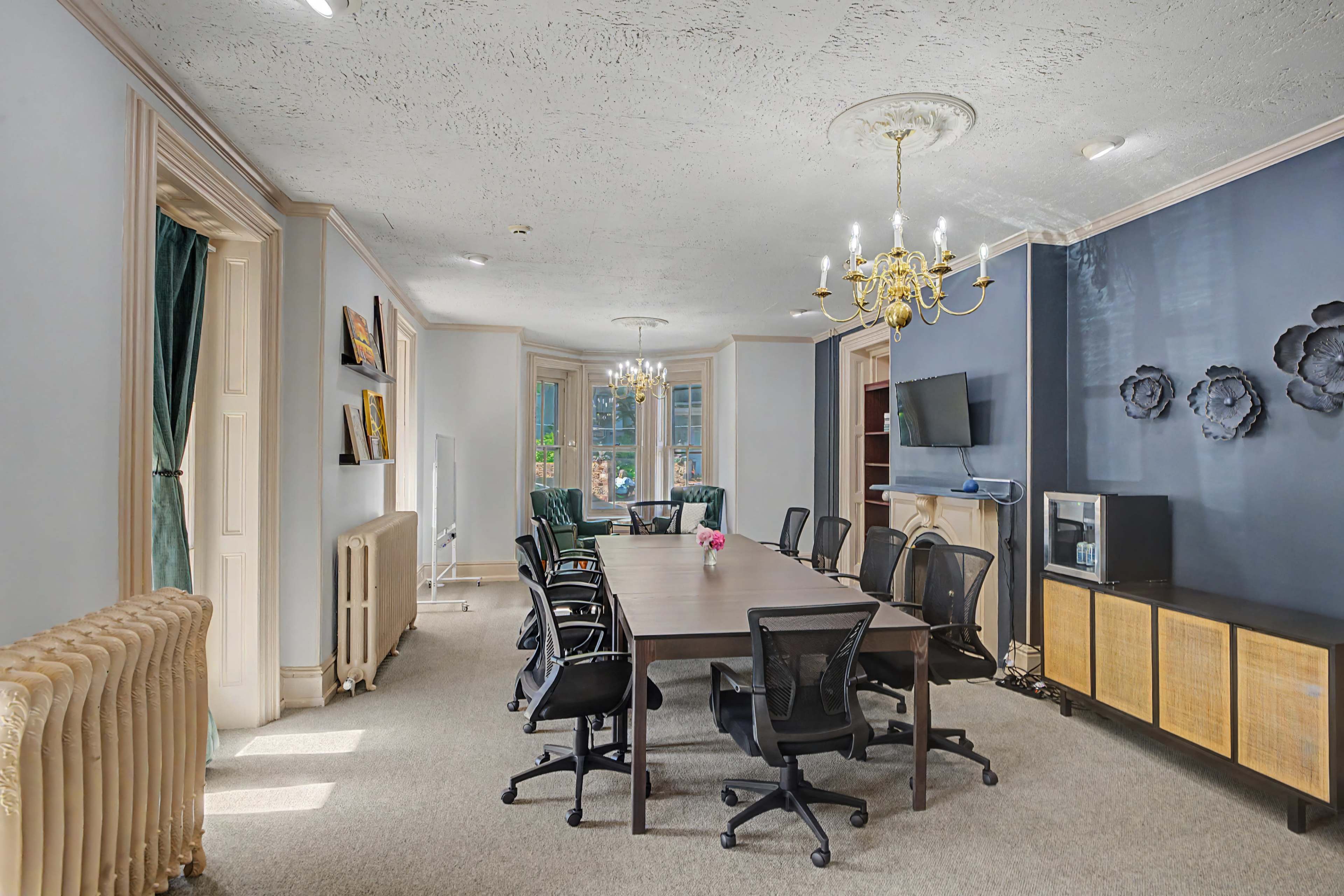 The image shows a spacious conference room with a large wooden table surrounded by black office chairs, a television mounted on the wall, and large windows allowing natural light to enter.