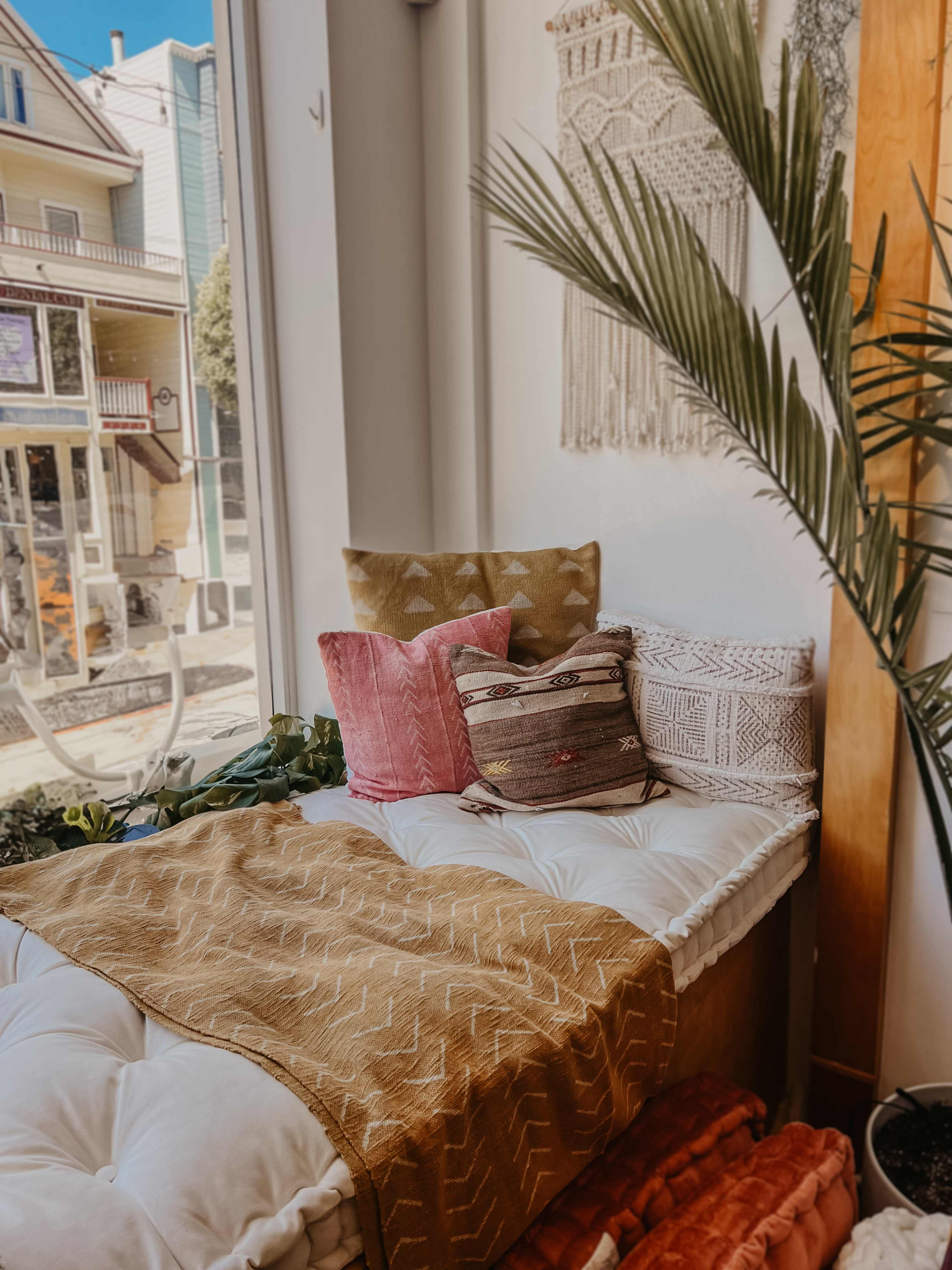 A cozy window nook is adorned with decorative pillows and a golden throw blanket, surrounded by plants and bright natural light.