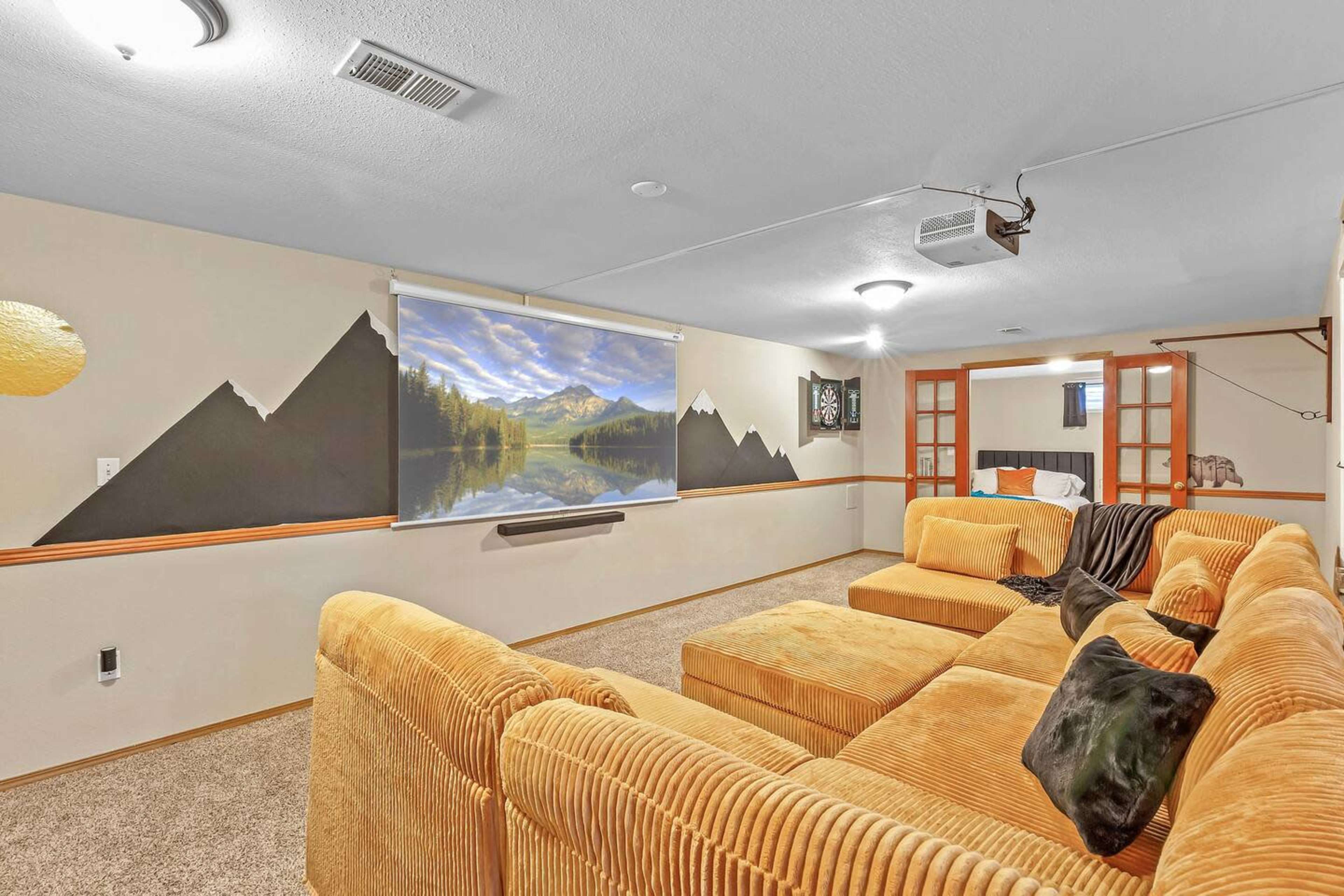 A basement living area with a large sectional sofa, a projector screen displaying a mountain scene, and painted mountain silhouettes on the wall.