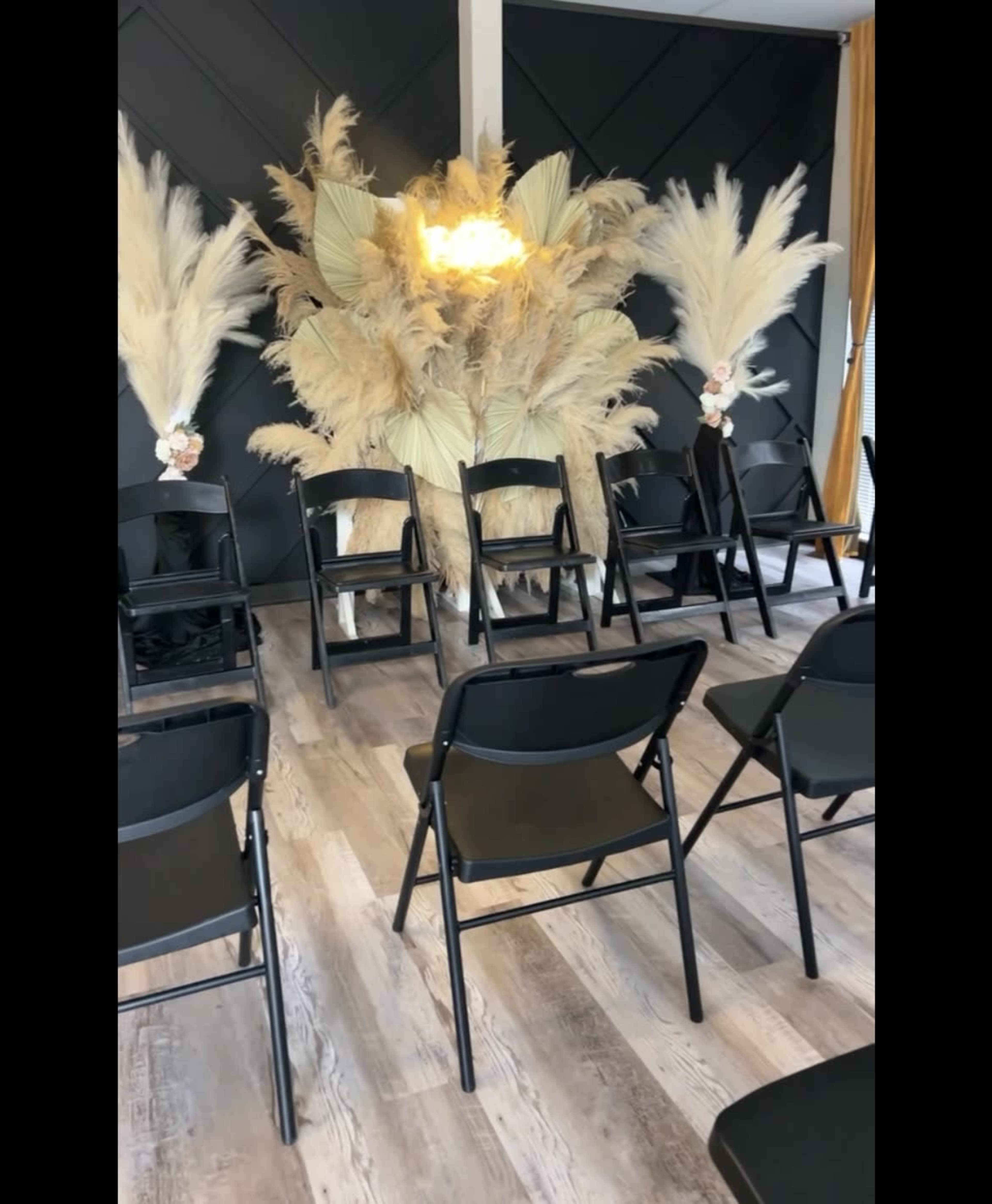 A set of black chairs is arranged in front of a large decorative backdrop made of pampas grass and a light fixture.