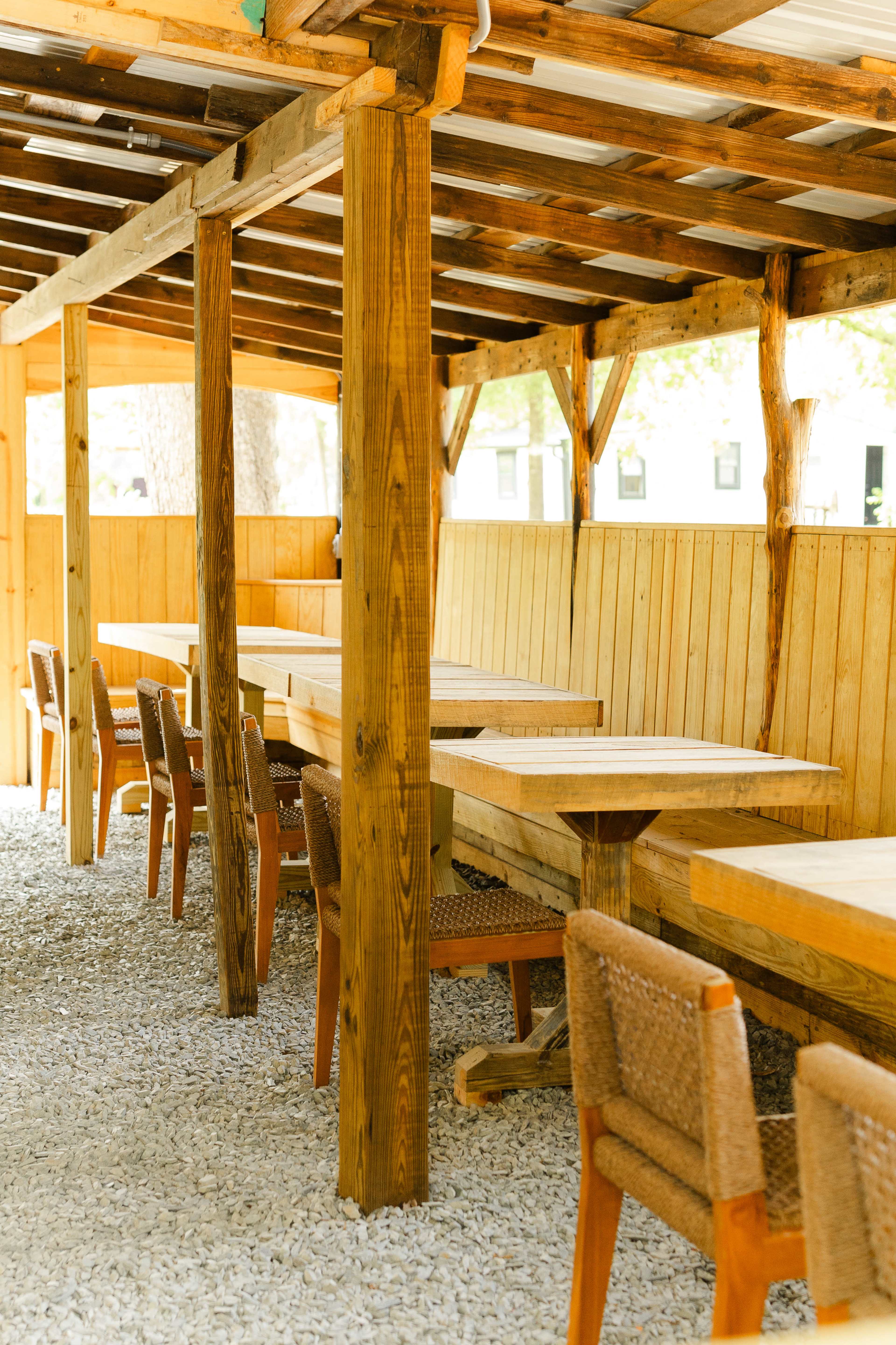 The image shows a rustic outdoor seating area with wooden tables and chairs arranged under a slatted roof.