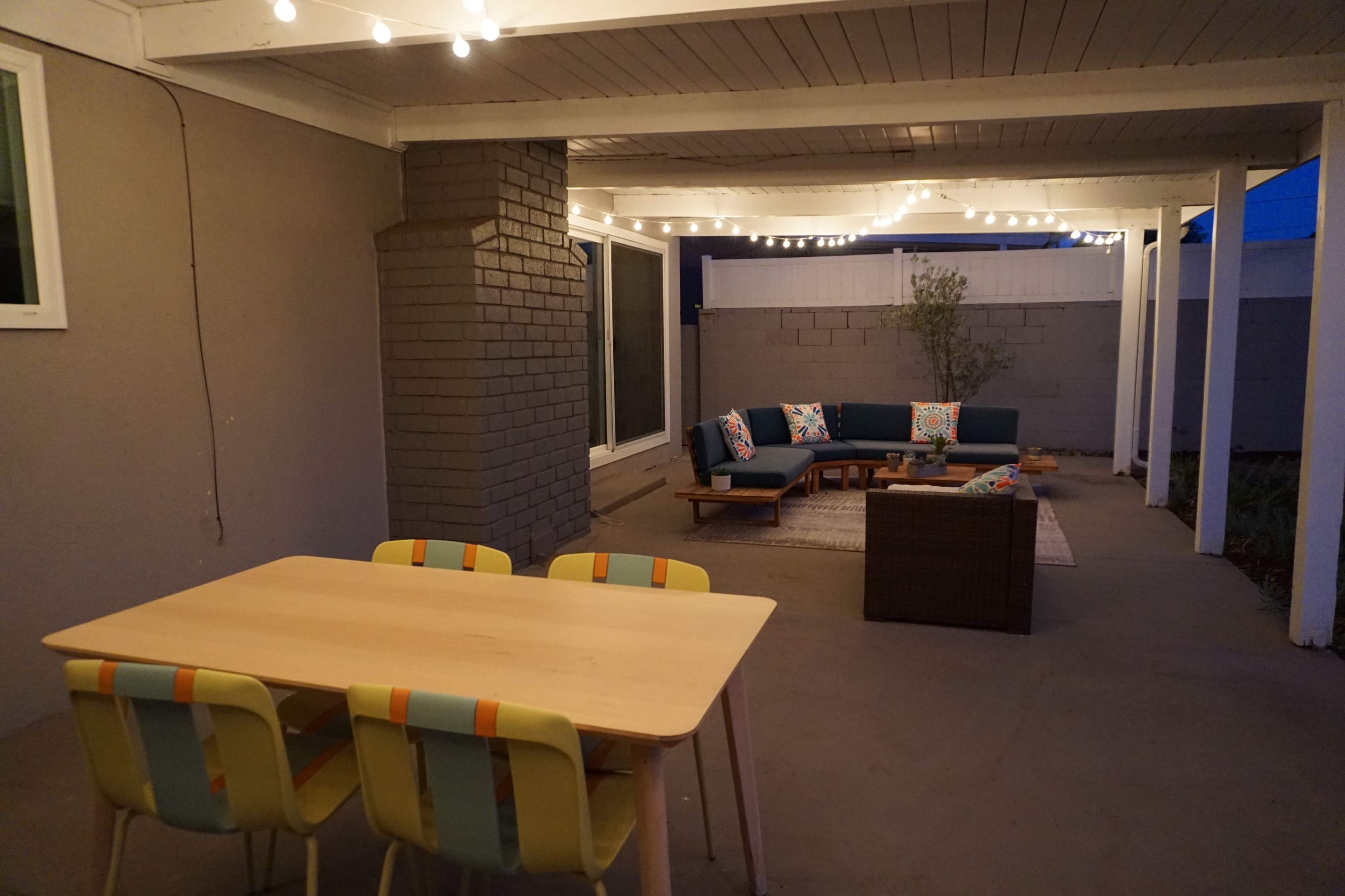The image shows a patio area featuring a seating arrangement with cushions, a coffee table, and a dining table with colorful chairs, all under a lit pergola.