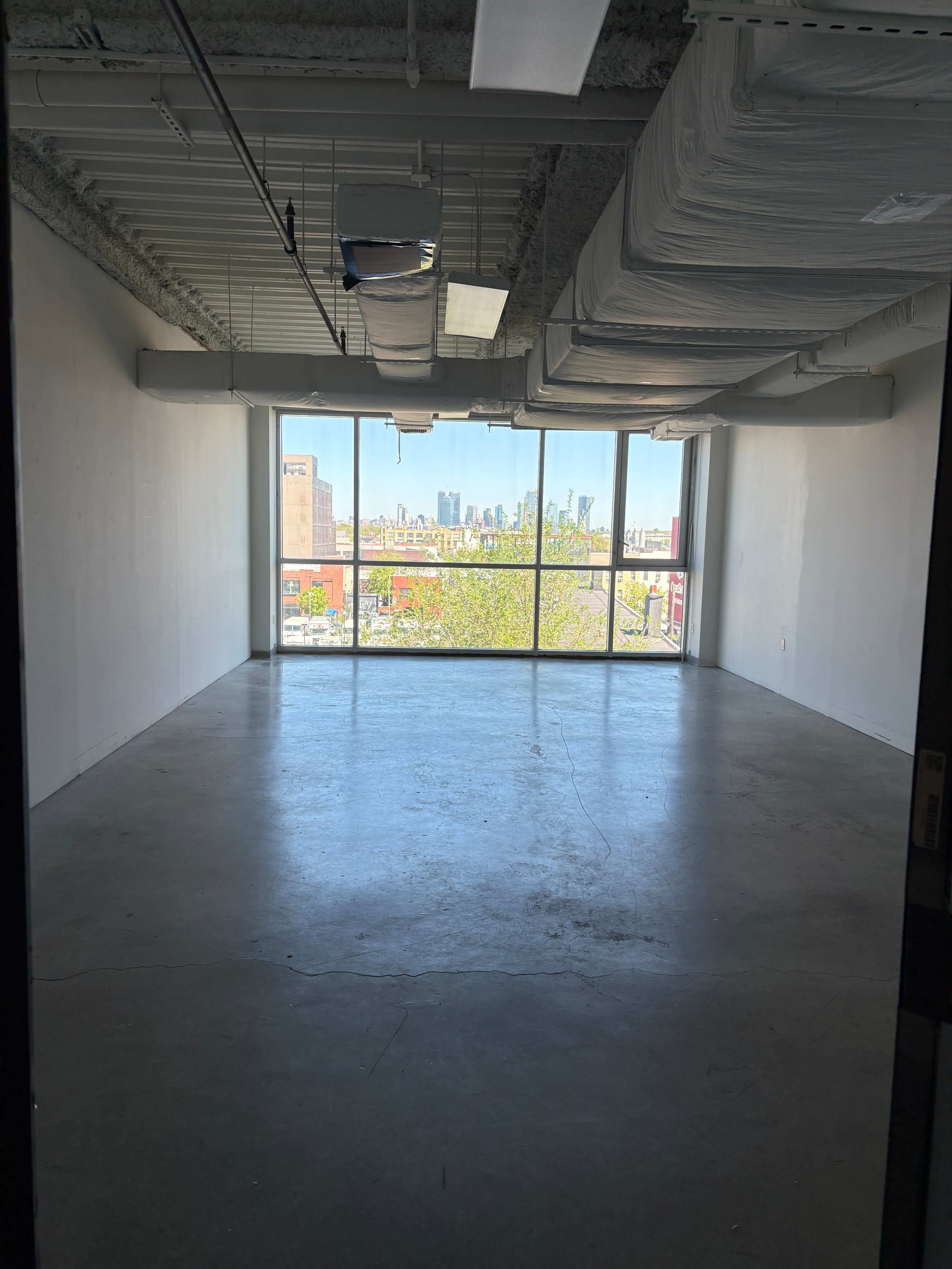A spacious, empty room with concrete flooring and large windows offering a view of a city skyline.